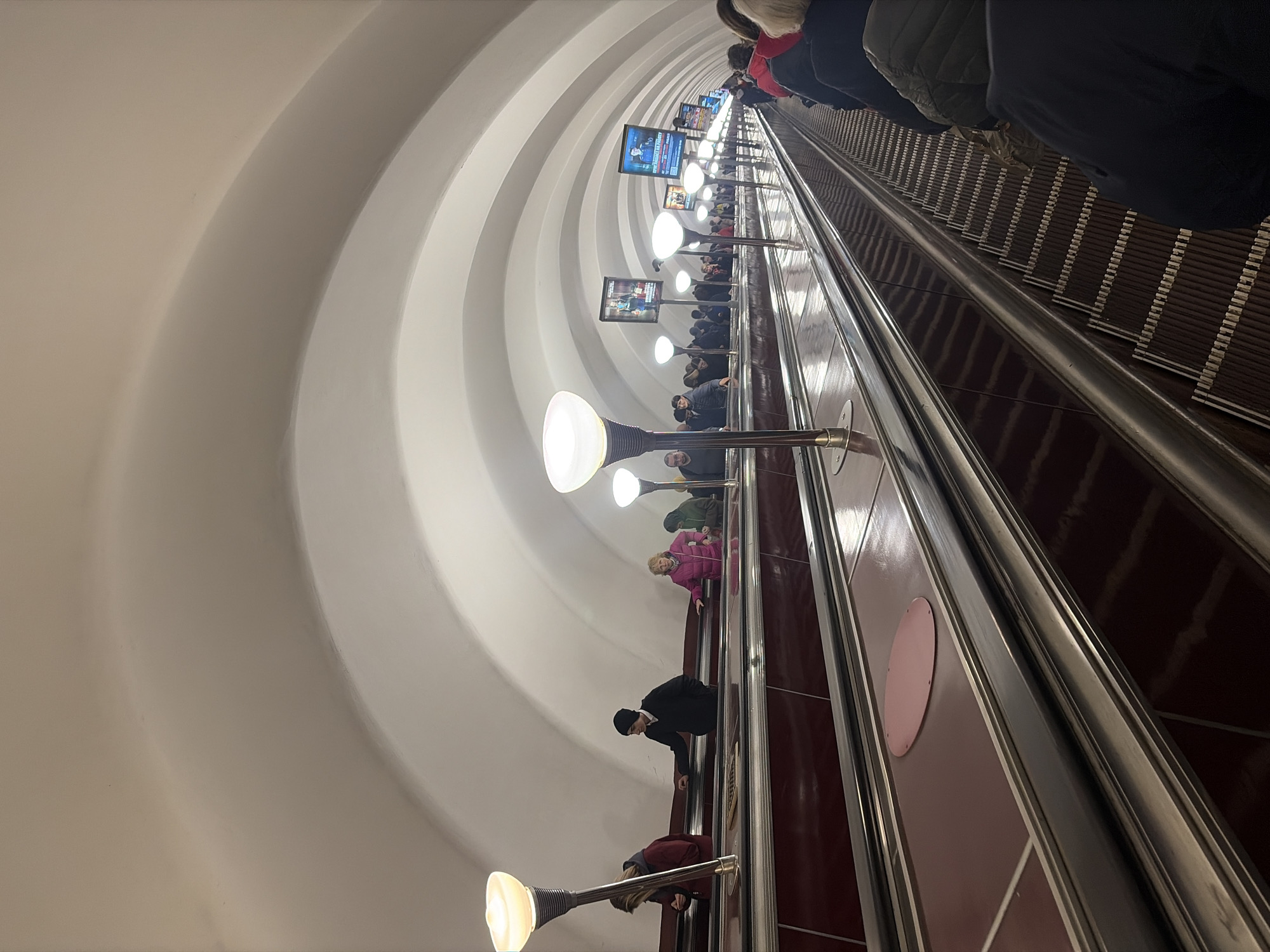This image shows an upward view of an escalator carrying people within the Saint Petersburg Metro in Russia. The escalator is flanked by walls with a series of arched recesses, illuminated by regularly spaced light fixtures. Advertisements or informational displays are positioned at intervals above the passengers. People are standing on the escalator, generally facing upward. The escalator's steps are visible, as are the metal handrails. The color scheme is primarily composed of whites, browns, and metallic silvers.