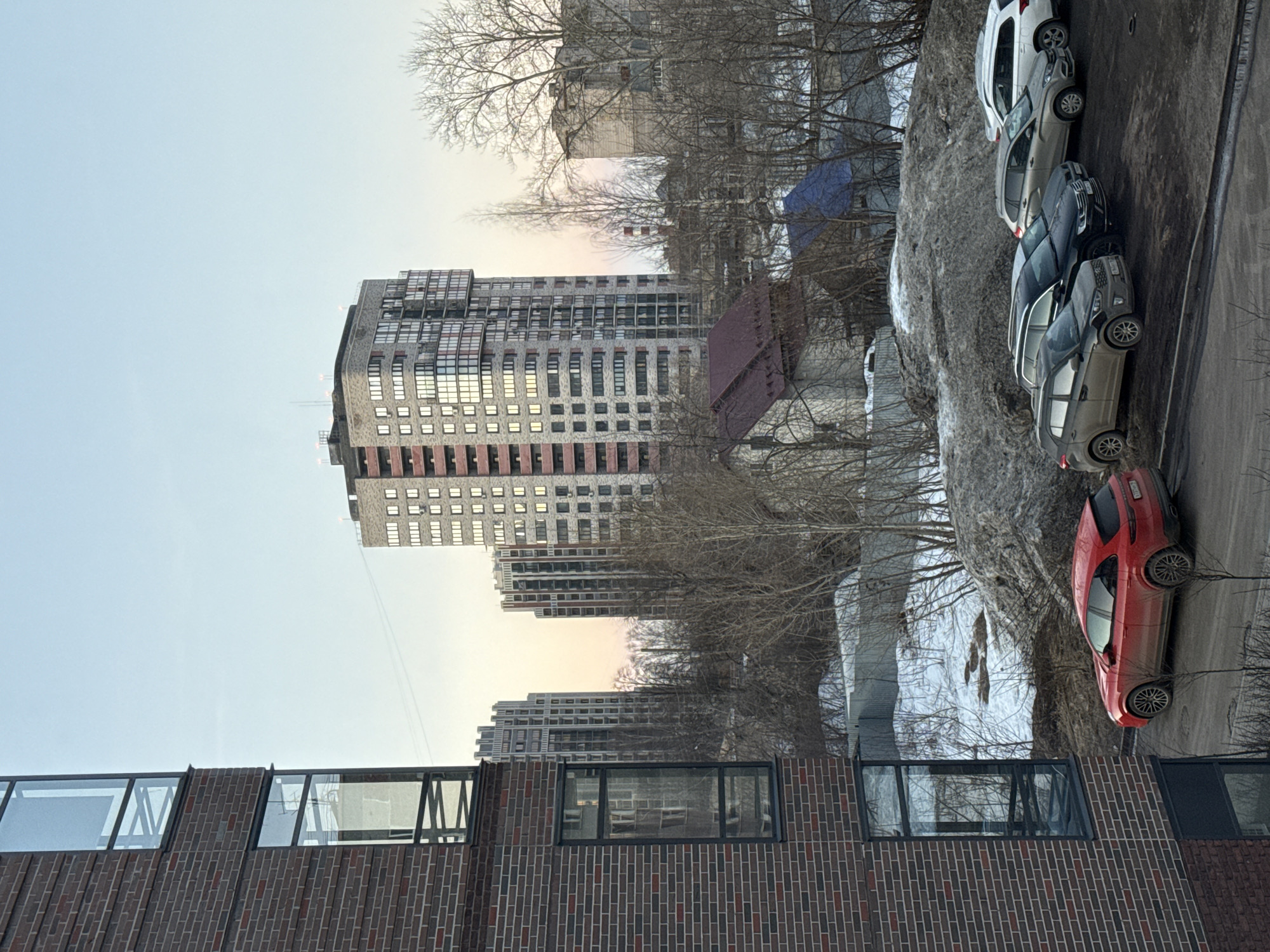 The image shows a view of Izhevsk, Russia, likely taken from an elevated position. In the foreground, a brick building with dark-framed windows is visible. Beyond this, a row of parked cars, including a red one, lines the street. Melting snow is visible next to the street. In the background, apartment buildings rise into the sky, partially obscured by bare trees. The sky is a pale blue-gray, suggesting early morning or late afternoon.
