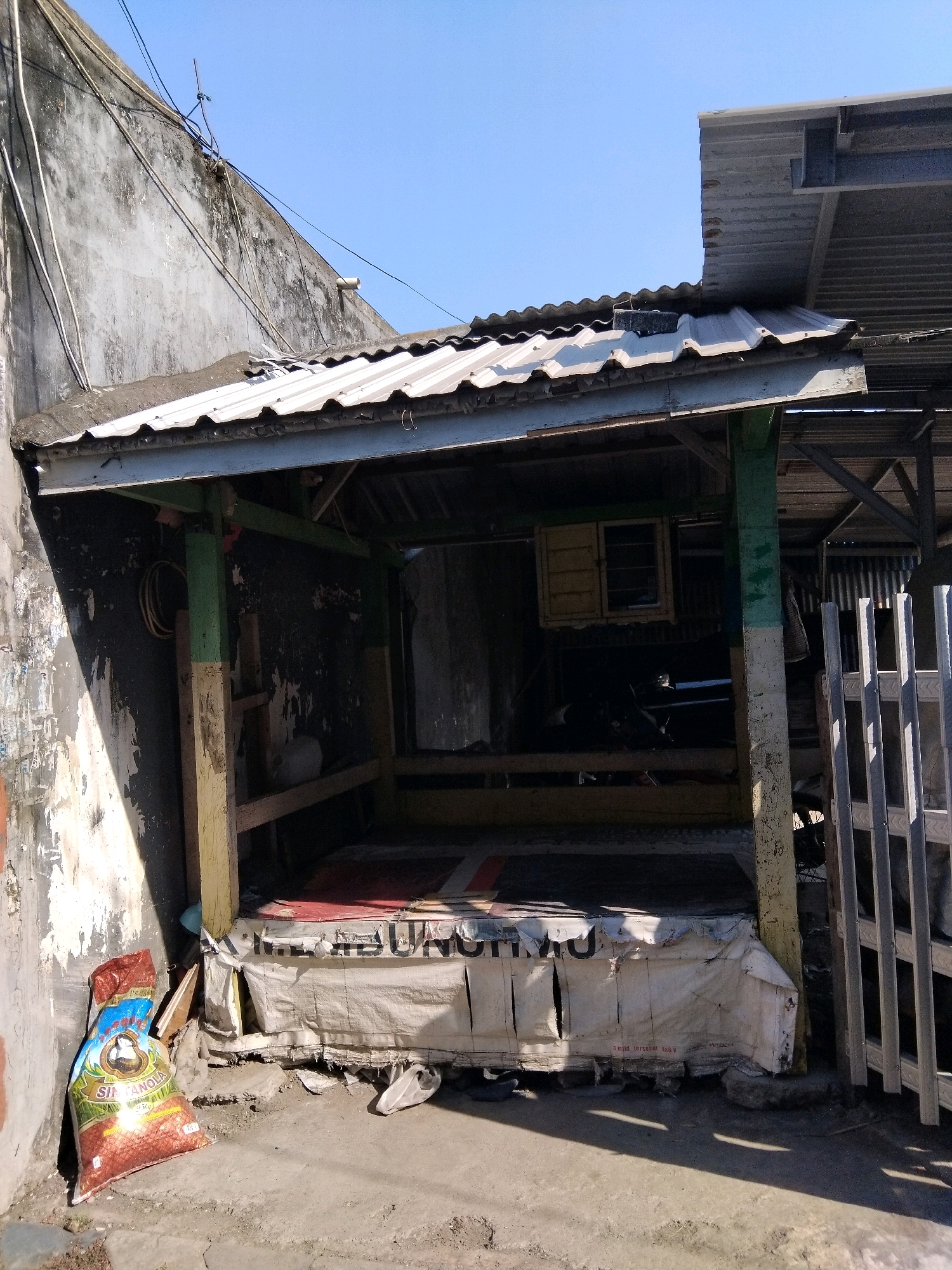 The image shows a weathered, open-air stall or shelter located outdoors in Kepuh Kiriman, Indonesia. The structure appears to be made of salvaged materials, with a corrugated metal roof supported by wooden beams, some of which are painted green and yellow. The walls of the stall are dark and appear to be made of rough-sawn wood or a similar material. In the foreground, a bag of what appears to be rice or grain, labeled "SANTANOLA," rests against a textured, possibly concrete or stucco, wall. There is a wooden fence or gate on the right side of the frame. The ground is uneven concrete or dirt. The scene is lit by bright sunlight, casting distinct shadows. The overall impression is one of a utilitarian and somewhat neglected structure in a typical urban or semi-urban environment in Indonesia. There are no people visible in the image.