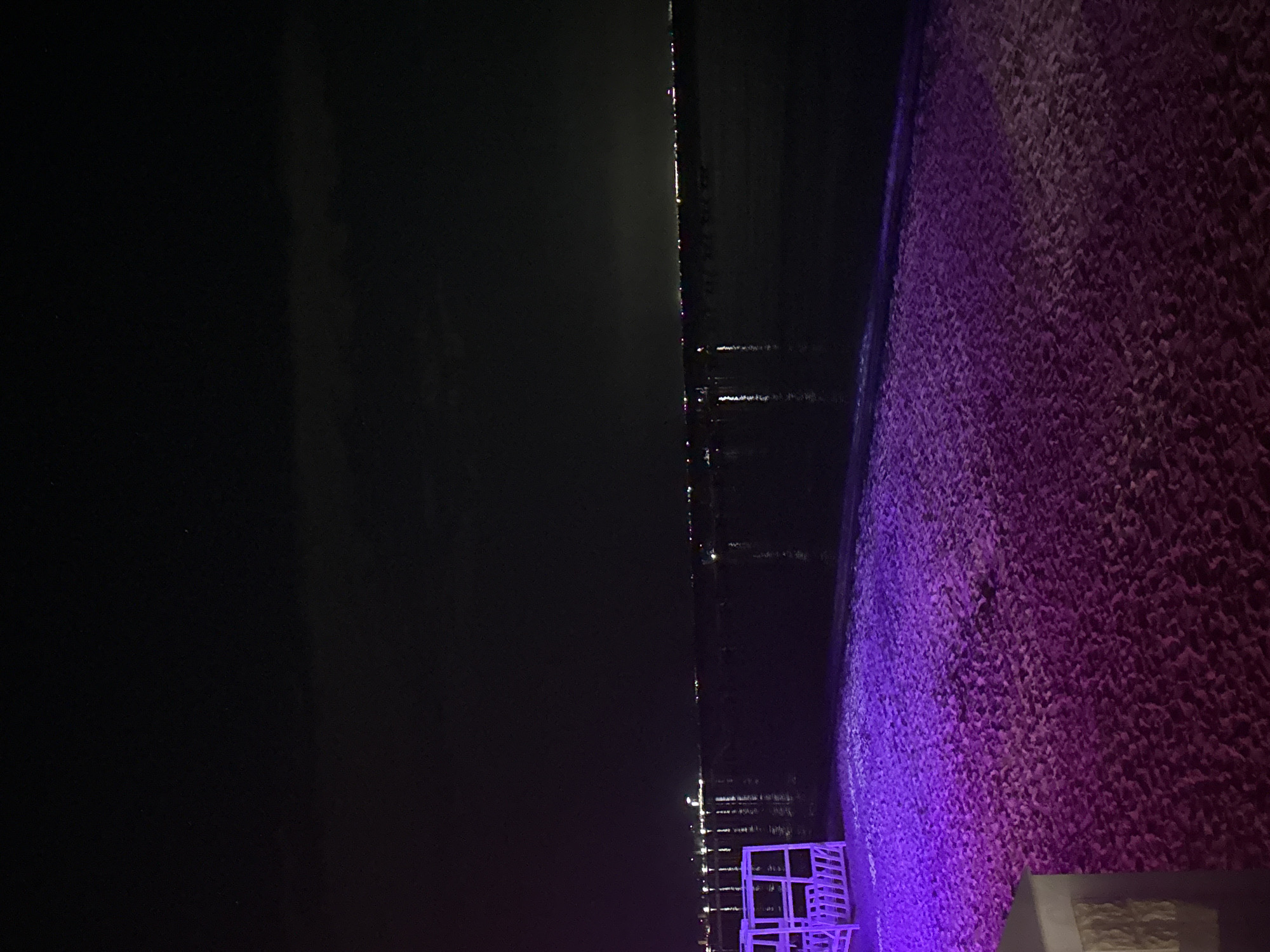 The image shows a nighttime view of the coastline in Yevpatoriya, Ukraine. The left side of the frame is dark, representing the night sky and possibly the sea. The right side features a wall or embankment illuminated with purple light. There are some chairs visible in the lower part of the frame. Lights from the city or distant structures are visible across the water.