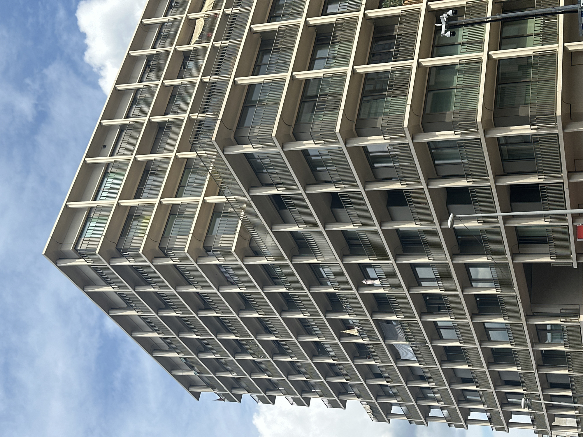 The image shows a tall, modern apartment building with a repetitive facade of balconies and windows. The building is made of concrete with dark metal railings on the balconies. There are a few items hanging from balconies, such as laundry or flags. The sky is blue with scattered white clouds. A street light with cameras is visible on the right side of the building. The architecture suggests a post-war or Brutalist style. The building appears to be located in an urban environment, potentially a residential area within the city of Amsterdam, Netherlands.