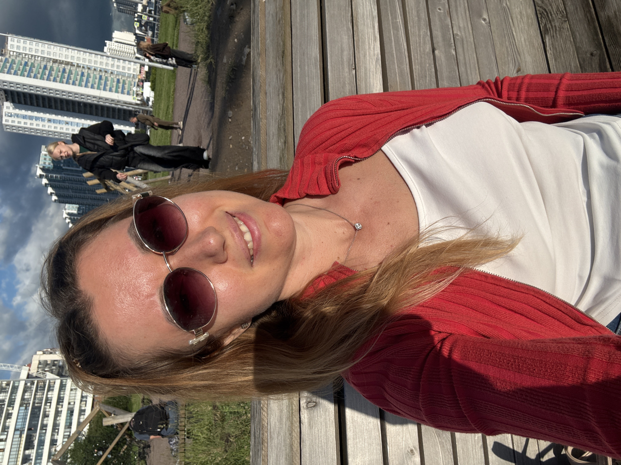 A woman with blonde hair and sunglasses sits on a wooden bench in an urban park. She wears a red zip-up jacket over a white top and a silver necklace with a diamond pendant. In the background, tall, modern buildings line the horizon under a cloudy sky. Other people are visible in the park, including a person in a black jacket and jeans walking, and a person with a backpack sitting on a low wall. The sun casts a shadow on the bench from the woman's left arm.