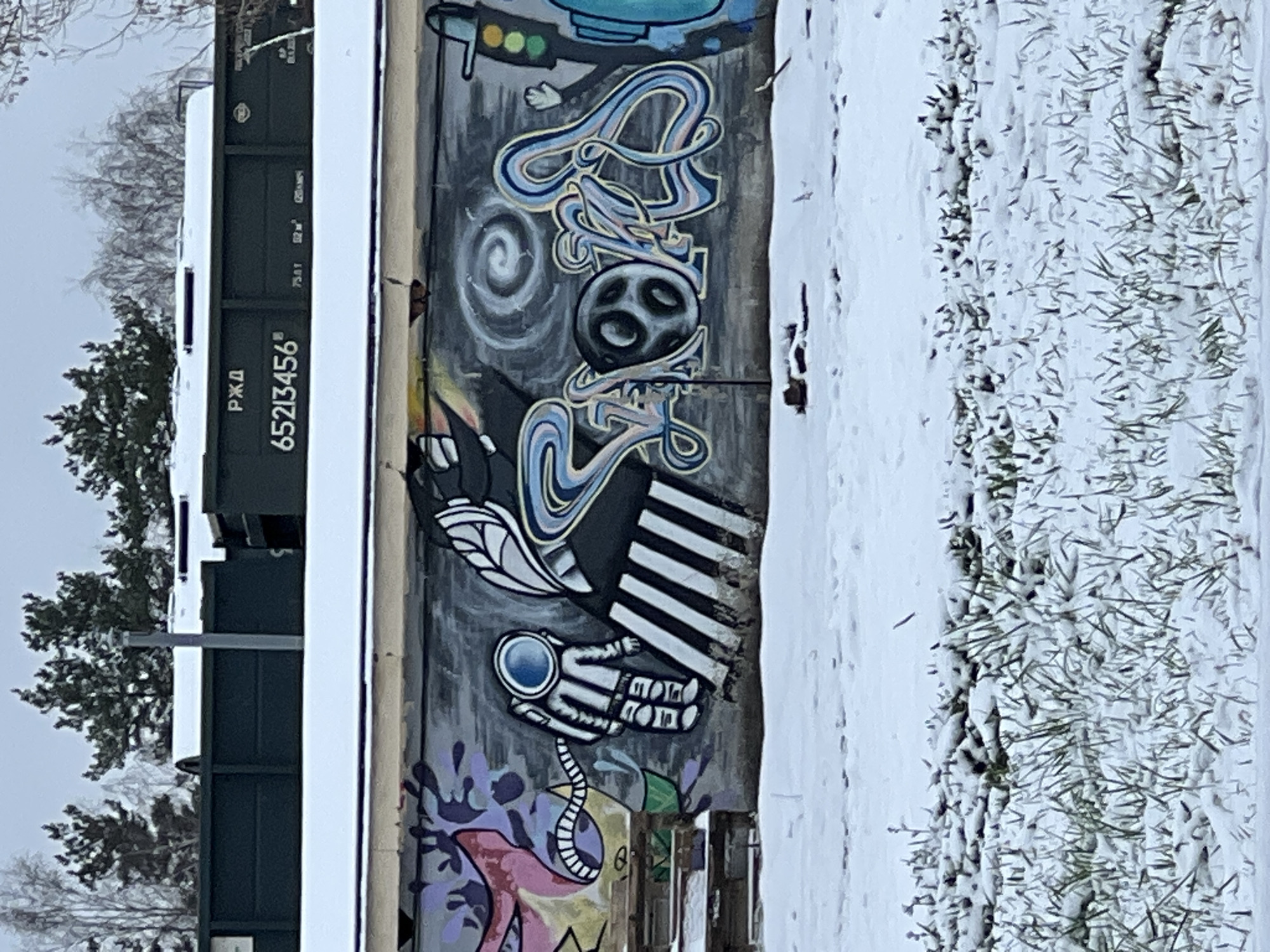 The image depicts a mural on a concrete wall in Chaikovsky, Russia, next to a section of railway tracks. The mural features an astronaut's gloved hand, a moon or similar sphere, and abstract designs. A train car is visible on the left side, with the markings "РЖД" and "65213456" visible on its side. The foreground is covered in snow-covered grass, suggesting a winter setting.