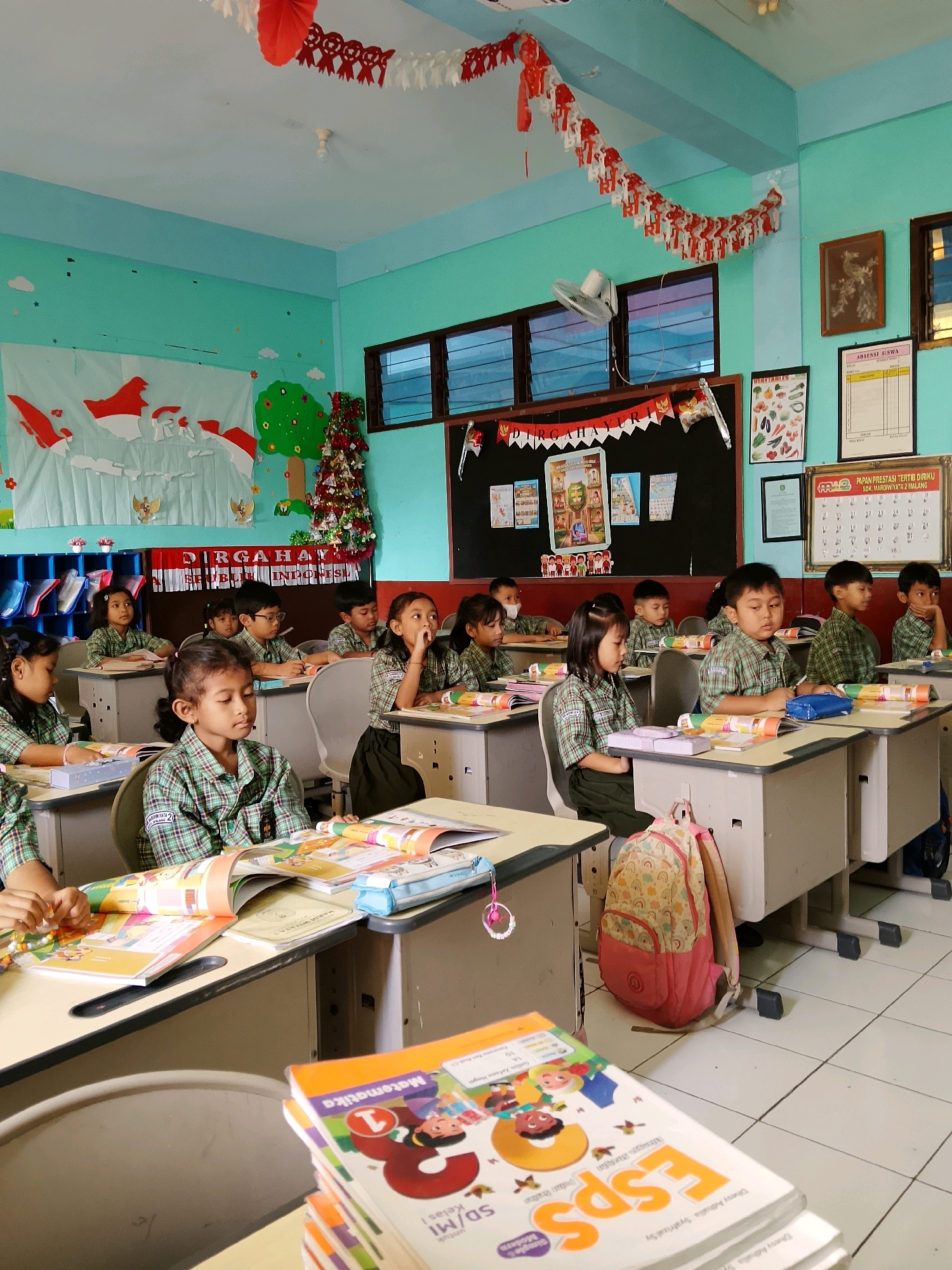 The image depicts a classroom in Malang, Indonesia, likely a primary school based on the young age of the students. The room is decorated with Indonesian national colors, red and white, with banners and garlands adorning the walls and ceiling. Several young students, boys and girls, are seated at desks, wearing matching green and white checkered uniforms. They appear to be engaged in learning activities, with books and writing materials on their desks. Some students are looking forward, perhaps at a teacher or the front of the room, while others are focused on their work. The classroom environment suggests a typical school day.