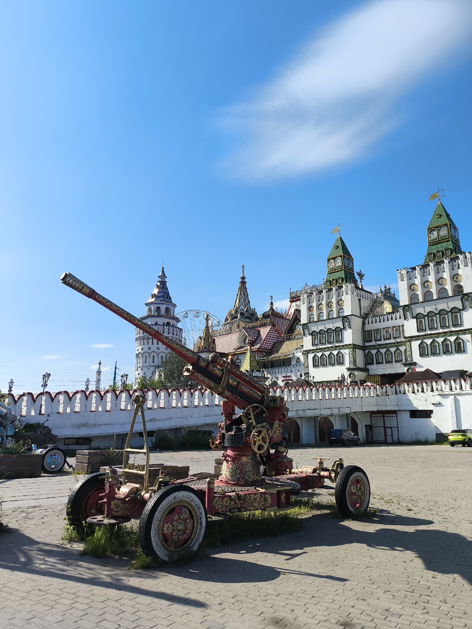 The image depicts an ornate, antique-style cannon displayed on a paved courtyard in front of a white walled fortress-like structure with green-topped towers. The cannon is predominantly red and gold, featuring intricate decorative patterns. Behind the cannon, the fortress exhibits a vibrant architectural style, with colorful roofs and numerous windows. A Ferris wheel is visible in the distance, behind one of the towers. The sky is a clear blue with a few wispy white clouds. The scene appears to be an outdoor exhibition or historical site.