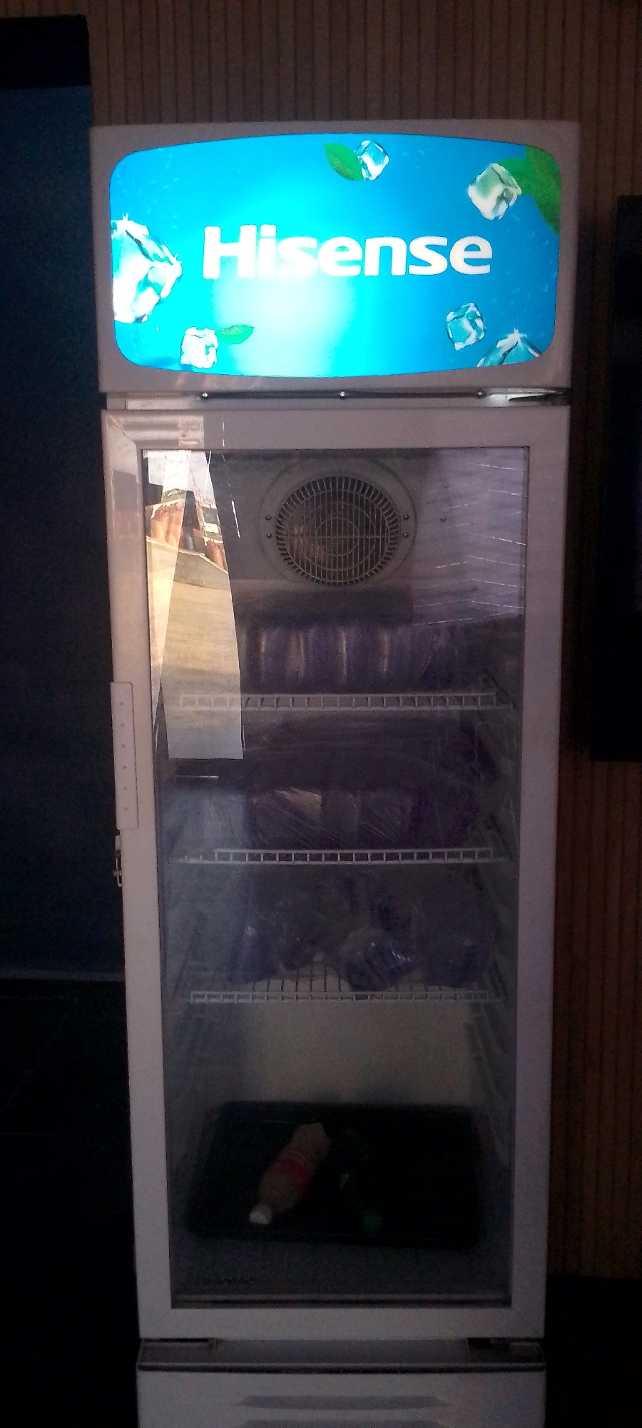 A full shot captures a Hisense brand refrigerator, its glass door slightly smudged, revealing shelves stocked with products wrapped in plastic. The top of the refrigerator features a lit panel with the Hisense logo, accompanied by graphics of ice cubes and mint leaves against a blue background. A circular fan is visible behind the glass door on the upper shelf. The bottom shelf holds a black tray containing two bottles, one with a red and white label, and another green bottle. The refrigerator is positioned against a background with vertical wooden paneling on the right and a dark, textured wall on the left. The lighting is dim, with the refrigerator's illuminated panel being the brightest element.