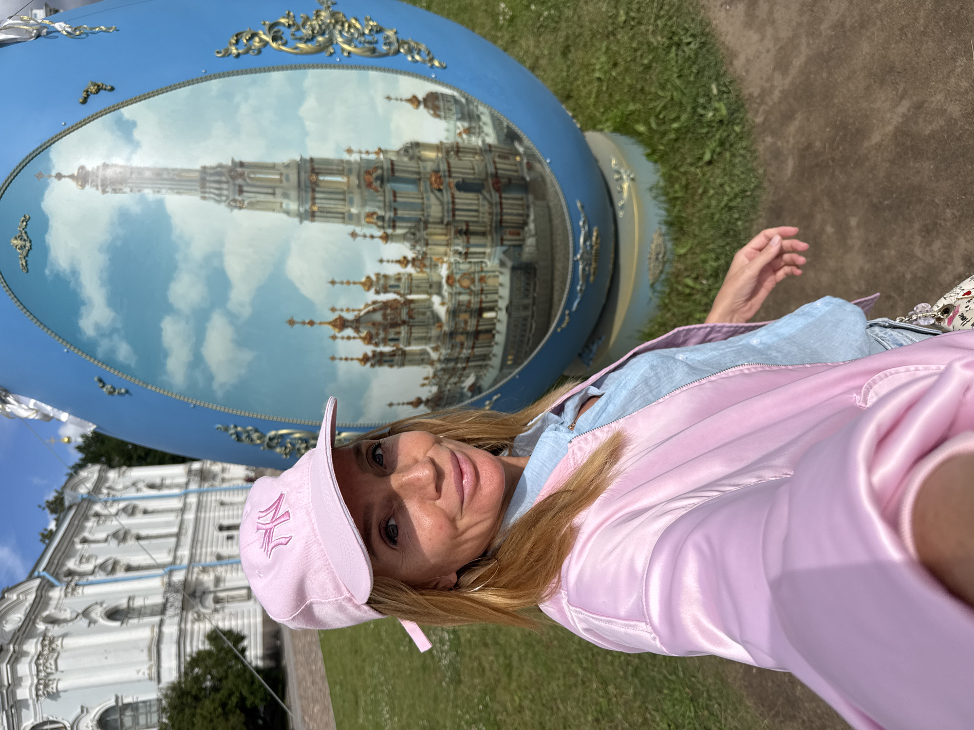 A woman in a pink baseball cap and a pink satin bomber jacket takes a selfie in front of a large blue egg-shaped sculpture with a painted image of a spire and ornate buildings. Behind the sculpture, a white building with classical architecture and trees are visible. The ground is covered in grass and dirt. The scene appears to be in the grounds of the Smolny Cathedral in Saint Petersburg, Russia, given the architectural style of the background building.