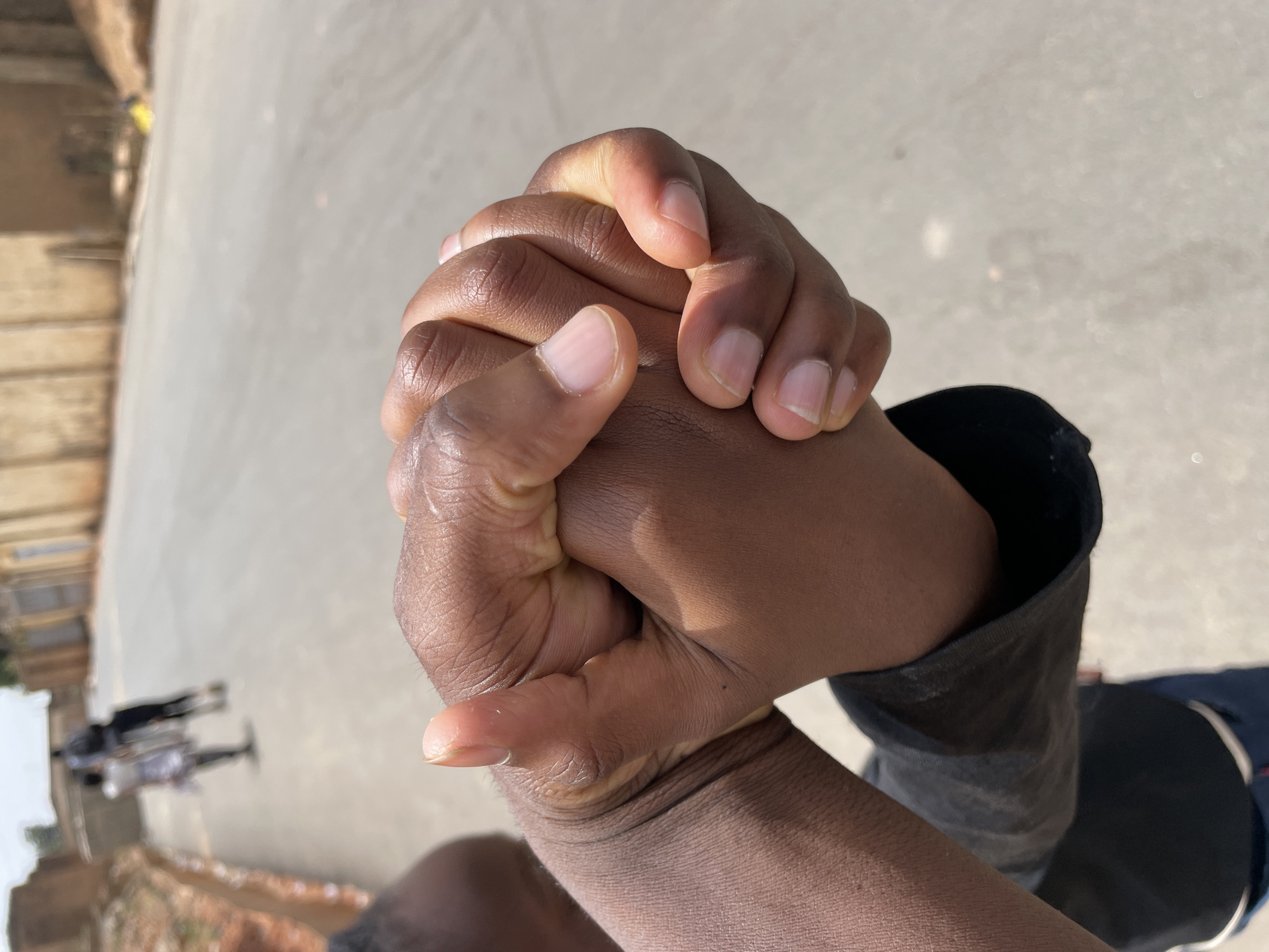 The image displays a close-up, high-angle view of two dark-toned hands tightly clasped together in the foreground, with fingers interlocked. The visible forearm on the left wears a dark long-sleeved garment. The background, largely out of focus, shows a light grey paved road or pathway receding into the distance in the city of Jos, Nigeria. Two blurry human figures are visible walking away from the camera on this road. To the right of the road, a low, light brown or tan wall or building facade runs along the roadside, while an earthen embankment, reddish-brown in color, is visible on the left. The scene is illuminated by natural daylight.
