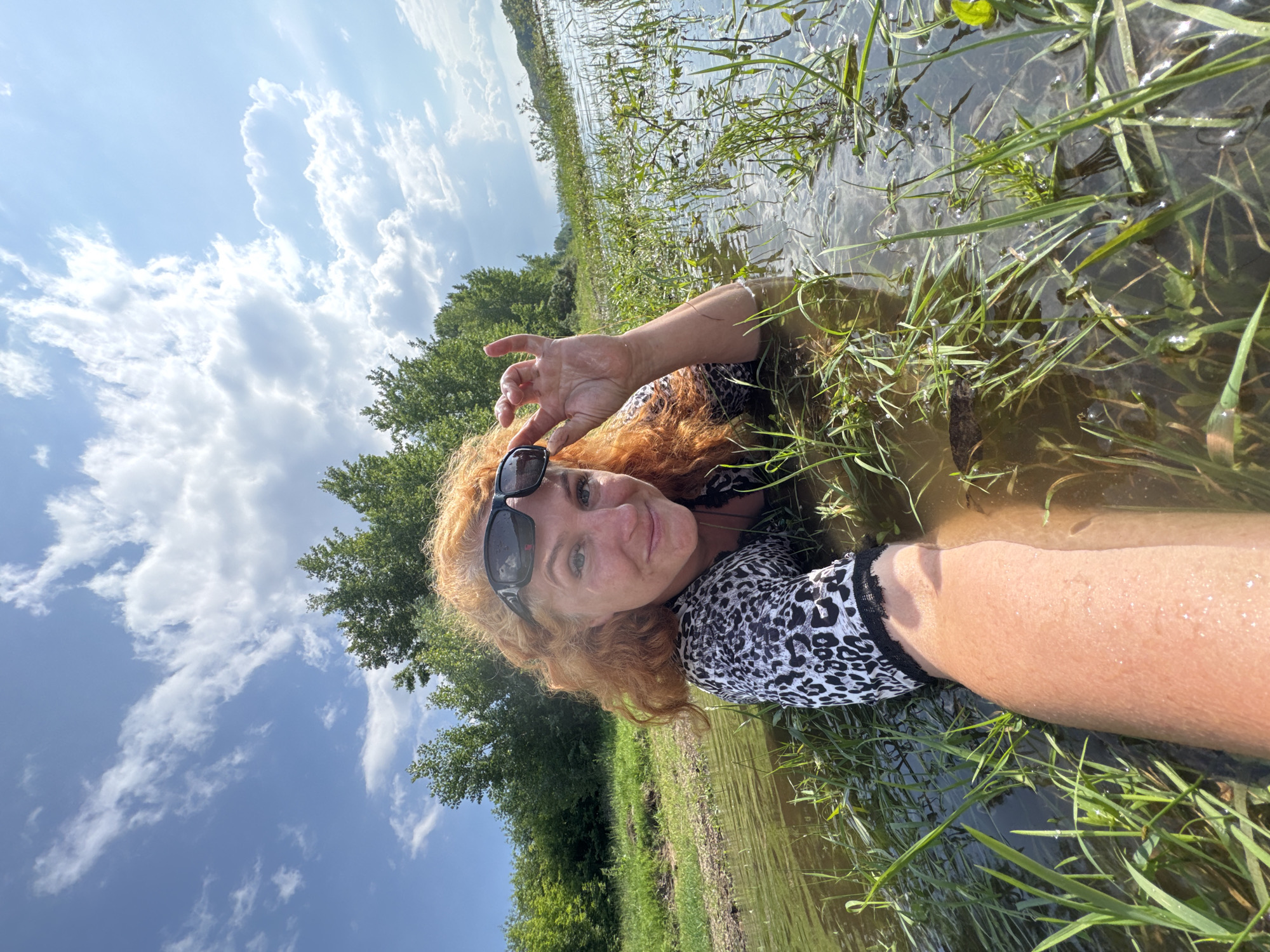 The image depicts a woman with red hair and sunglasses lying in shallow water surrounded by grass and greenery. She is wearing a black and white leopard print top. The background shows trees and a blue sky with clouds. The scene appears to be a natural outdoor environment, possibly a riverbank or a flooded area. There is no indication of the city of Chaikovsky, Russia, within the image.