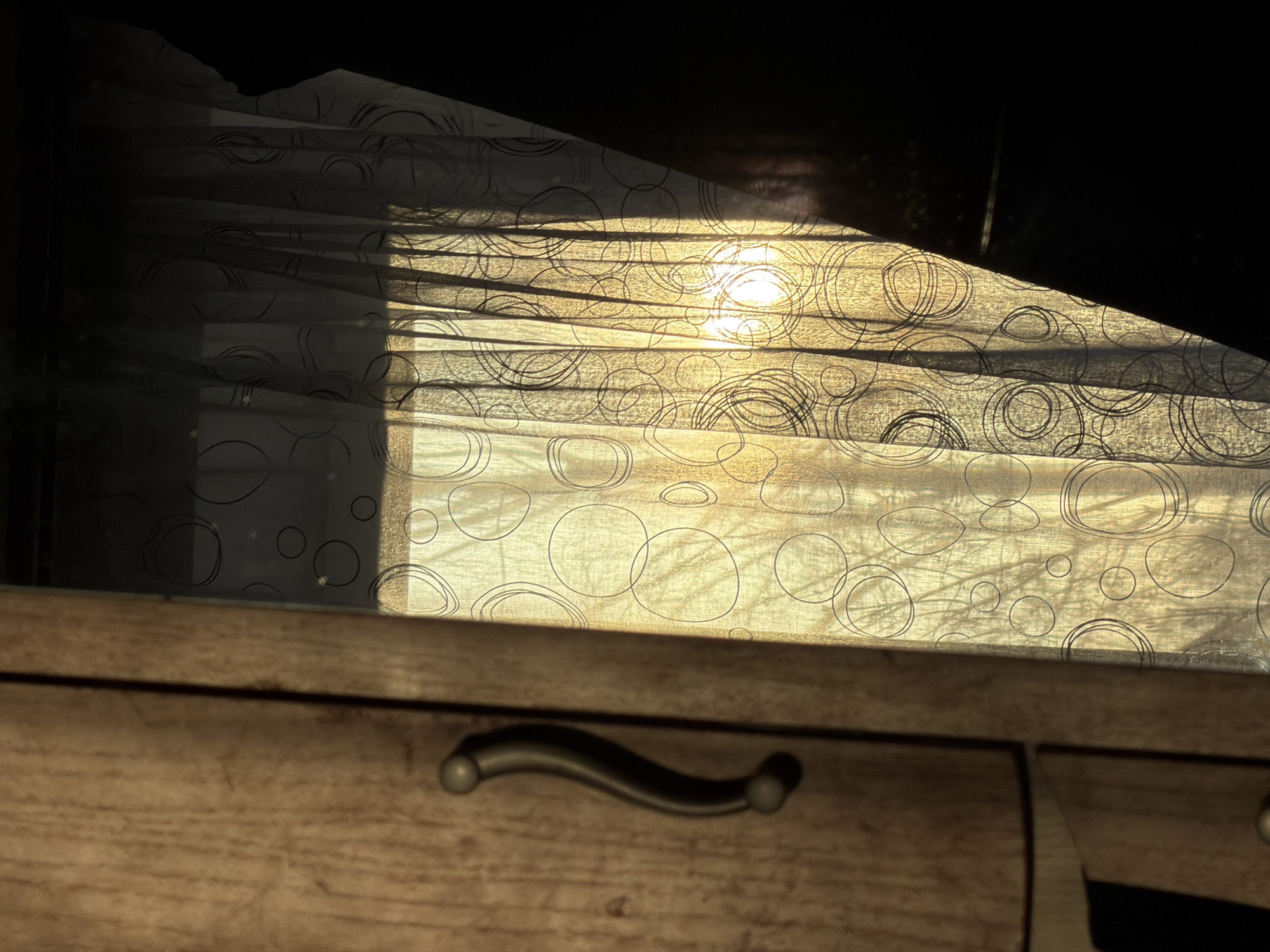 A low-angle indoor shot focuses on a section of a wooden dresser with a silver-colored, curved handle on one of its drawers. Above the dresser, a light-colored, patterned curtain hangs. The curtain has a repeating design of black circles. Sunlight filters through the curtain, casting a warm glow and creating shadows across the fabric. The top of the curtain appears torn or uneven. The background behind the curtain is dark and indistinct. There is no verifiable information in the image to confirm its location in Izhevsk, Russia, or any other specific location. No people are visible.