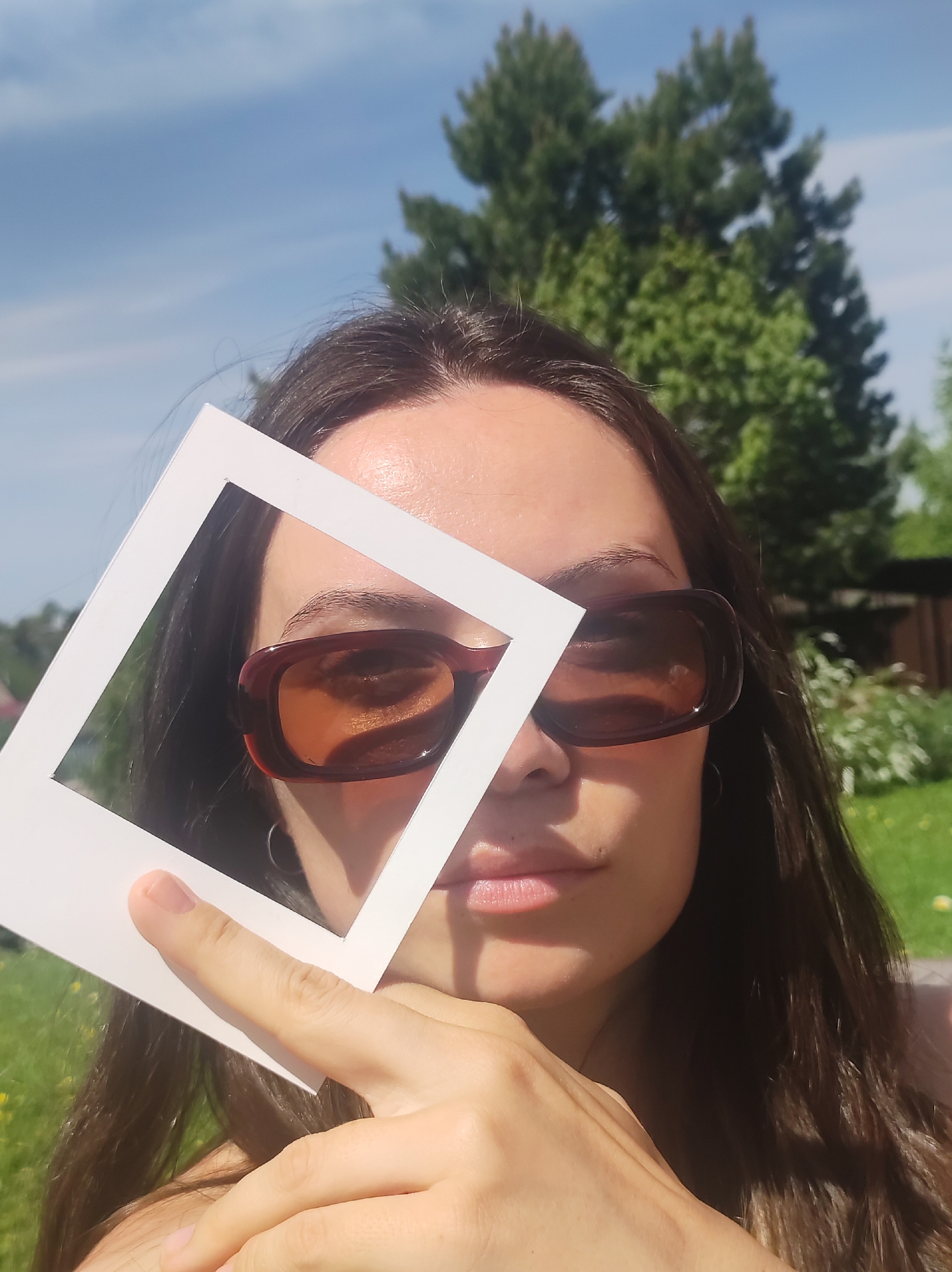 A woman with dark hair and reddish-brown sunglasses holds a white square frame up to her face in an outdoor setting. The frame partially covers her left eye. She rests her chin on her hand. A tree is in the background, partially obscured by the woman's head. The sky is visible in the top left corner. The location is a lawn in Фоки, Russia.