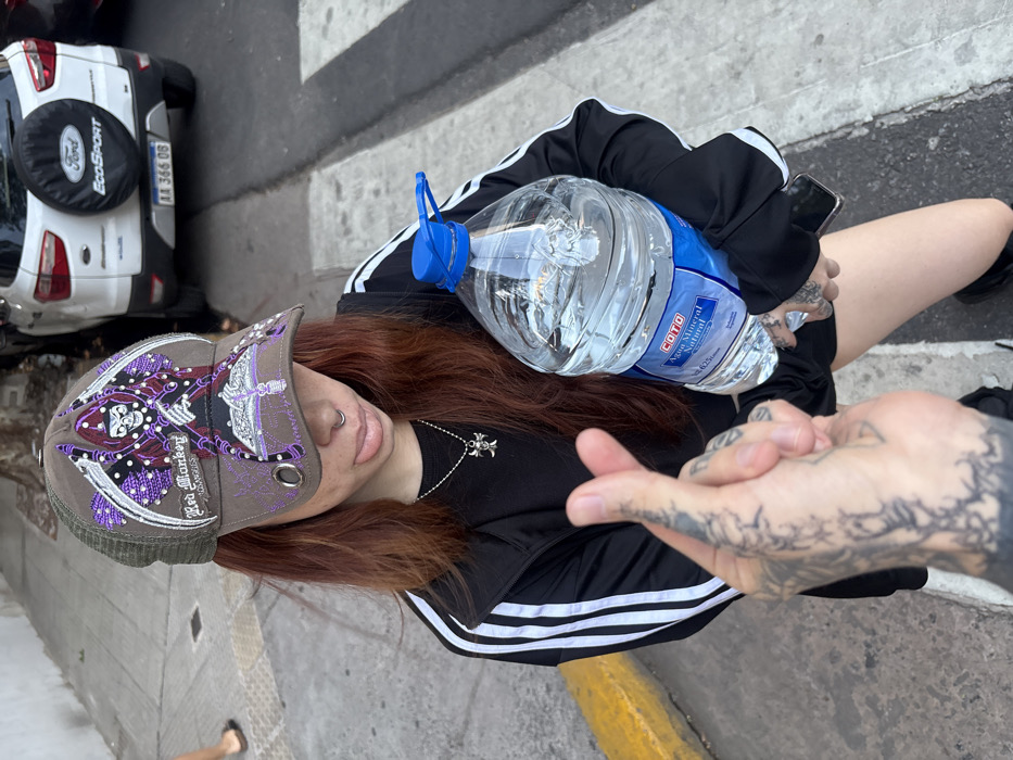A person with red-brown hair, a nose ring, and tattoos wears a decorated cap and black Adidas jacket, holding a large water bottle. Another tattooed hand is intertwined with theirs. A white Ford EcoSport is parked nearby, facing the same direction. They are standing on a street with a yellow curb and a zebra crossing.