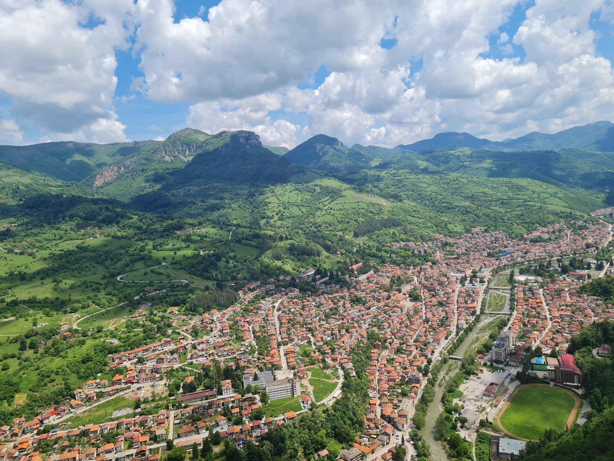 A high-angle view shows the town of Veliko Tarnovo, Bulgaria, nestled among the green hills and mountains under a partly cloudy sky. The town features closely packed buildings with predominantly red-tiled roofs. A river runs through the center of the town, dividing the urban area. A green sports field with a running track is visible along the riverbank. The landscape is dominated by lush green vegetation, indicative of a verdant environment. The clouds are puffy and white, scattered across the blue sky.