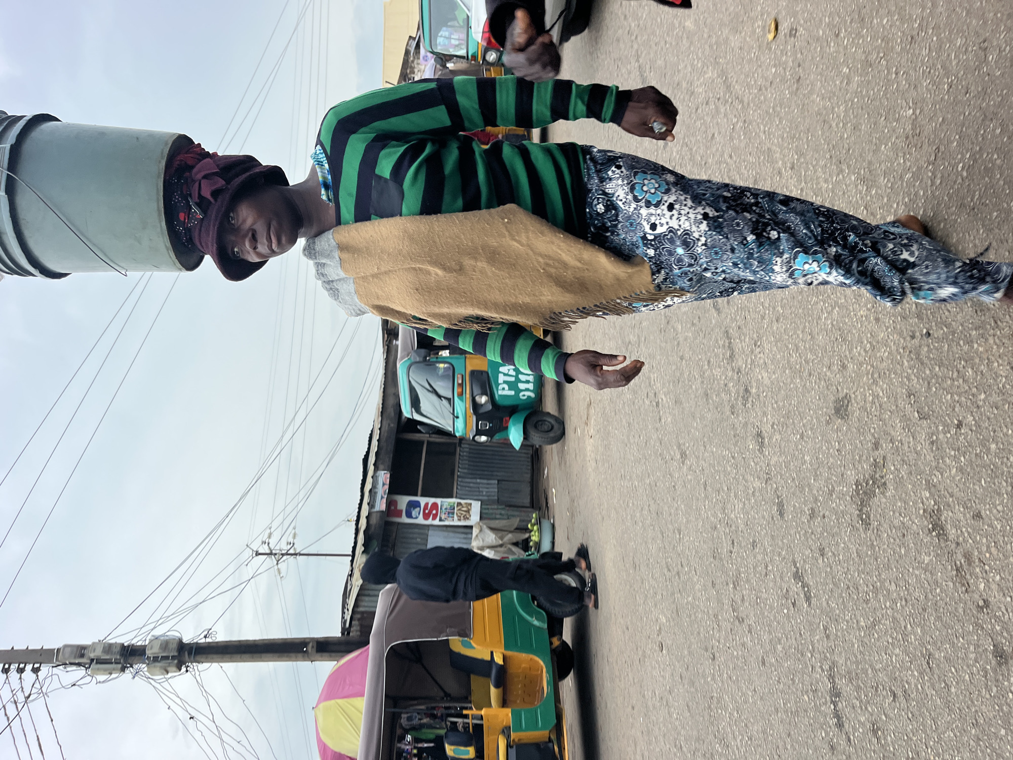 A woman walks on an asphalt street, balancing a large green bucket on her head. She is wearing a brown and maroon head wrap, a green and black striped long-sleeved shirt, a tan scarf draped over her shoulders, and a patterned blue and white skirt. The street is lined with buildings and various vehicles, including tuk-tuks. Overhead, there are numerous electrical wires and poles. The sky is overcast.