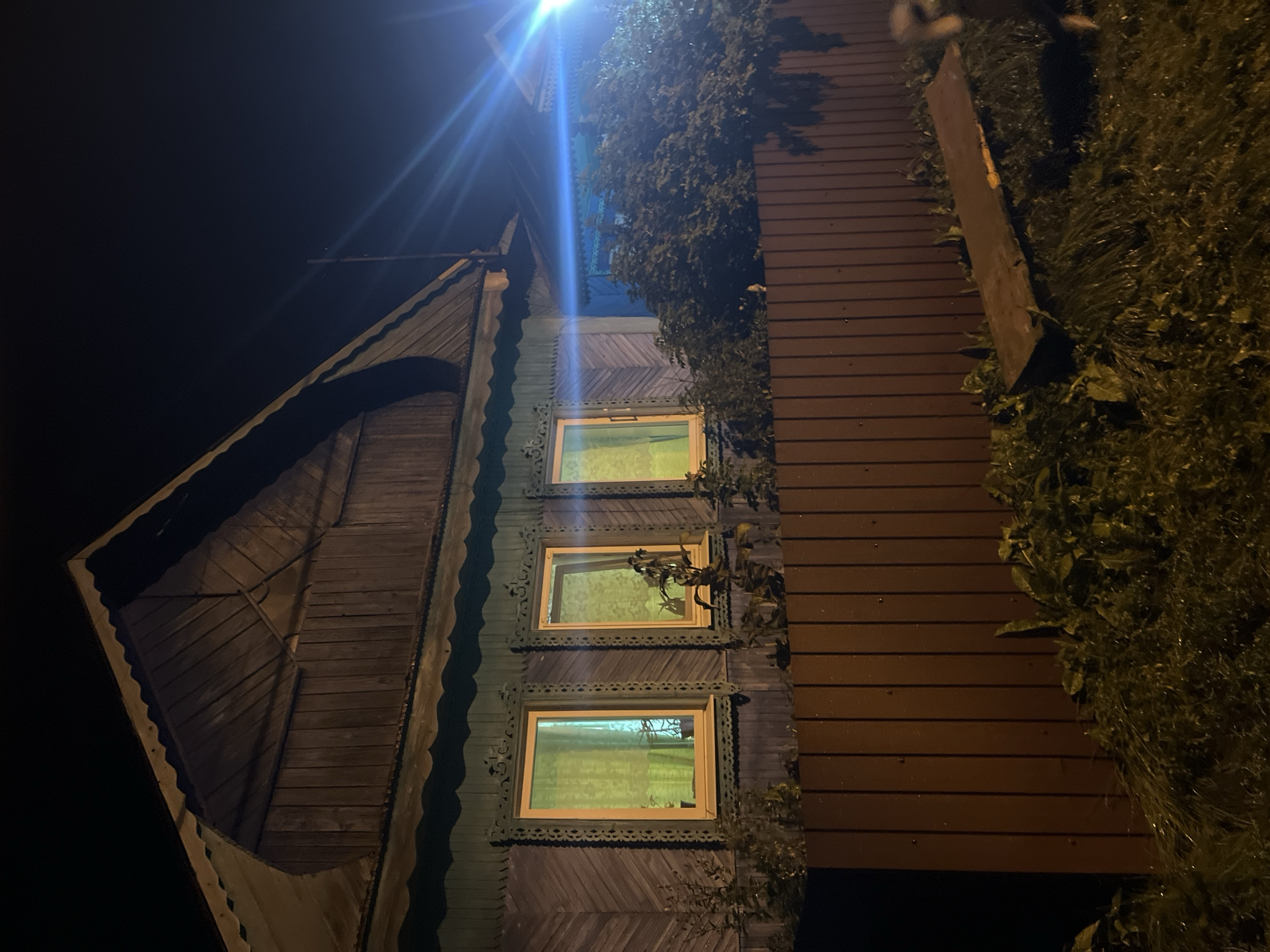 The image shows a wooden house at night in Nizhny Tagil, Russia. The house has a distinctive wooden facade with decorative carvings around the windows. Three windows are visible, all illuminated from within with a yellowish light, suggesting occupancy. A brown wooden fence runs across the foreground, partially obscuring the lower part of the house. Beyond the fence, there is overgrown vegetation and a makeshift wooden bench. A bright, blue light source is visible in the upper right portion of the image, casting lens flares and illuminating the scene. The overall atmosphere is dark and somewhat eerie, typical of a nighttime rural or semi-rural setting.