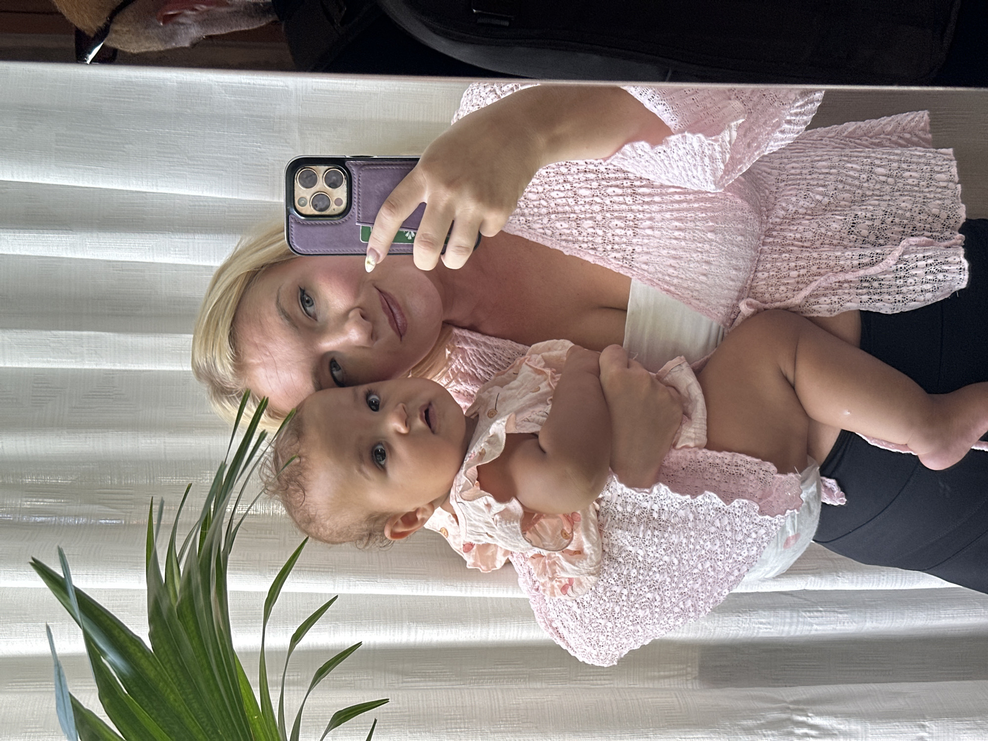 A blonde woman holds a baby in her arms, taking a selfie in a mirror. The woman is wearing a pink, open-knit cardigan over a white strapless top and black leggings. The baby is dressed in a light pink ruffled romper and is looking towards the camera. A plant with large green leaves is visible on the left side of the frame. The background consists of white curtains. The scene appears to be indoors.