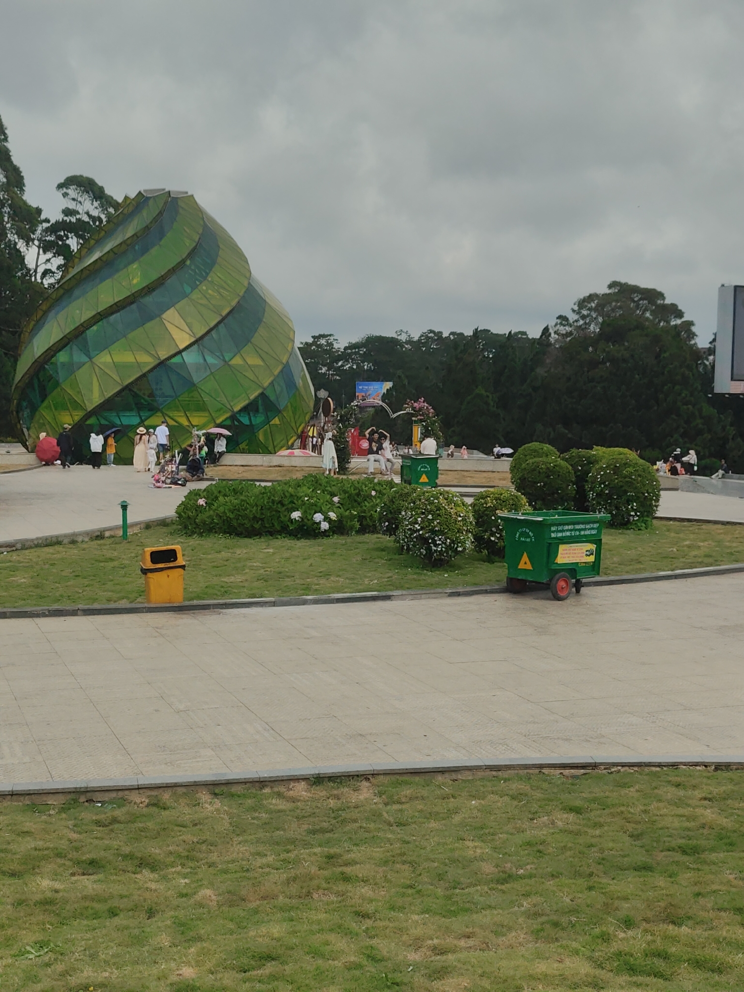 The image depicts a public square on an overcast day in Da Lat, Vietnam, centered around the distinctive Artichoke Bud structure within Lam Vien Square. The dominant architectural feature is a large, spiraling, multi-faceted glass building, primarily green and yellow, located in the mid-ground on the left. The sky above is a uniform light grey, indicating cloud cover.
Numerous people are gathered around the base of the glass structure and dispersed across the paved square. Several individuals are holding open umbrellas in various colors, including pink, red, and white. Groups of people are standing and walking, with some appearing to congregate near the building. On the far right in the background, a large screen or billboard is partially visible amidst dense green trees that line the perimeter of the square.
The ground consists of extensive light grey paved areas patterned with rectangular tiles, interspersed with segments of green grass. Several manicured green bushes, some adorned with small white flowers, are planted in beds within the paved sections. In the left mid-ground, a yellow rectangular trash bin with a black lid rests on the grass. To the right, on the paved area, a green two-wheeled utility cart with red wheels is visible. The cart bears white Vietnamese text, including "HÃY GIỮ GÌN MÔI TRƯỜNG SẠCH ĐẸP" (Please keep the environment clean and beautiful), "THU GOM RÁC TỪ 5H - 9H HẰNG NGÀY" (Collect trash from 5 AM - 9 PM daily), and "ĐÀ LẠT" with a city logo. A small green bollard is positioned near the yellow trash bin. Further back, partially obscured by bushes, another smaller green trash bin is present.