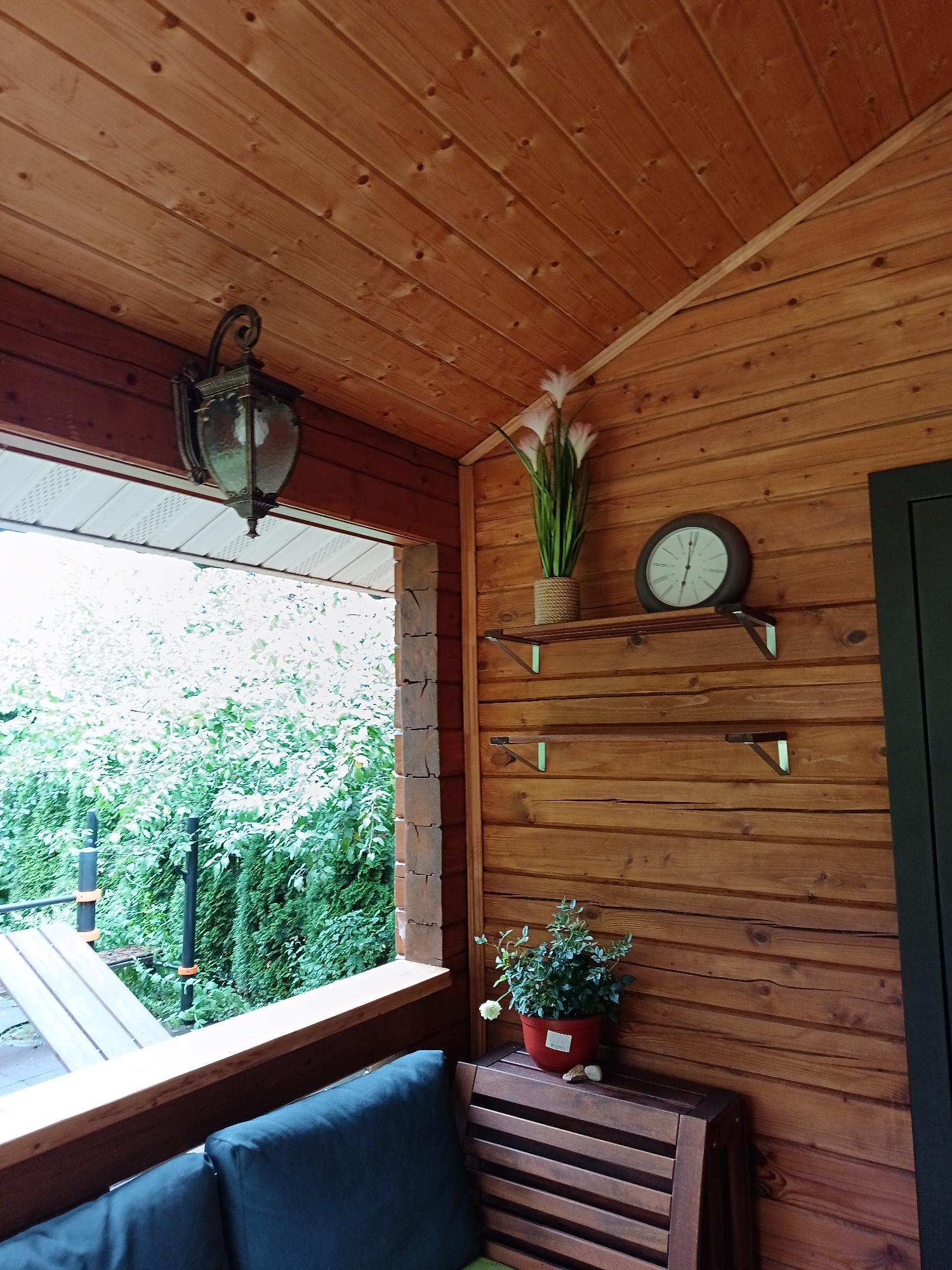 The image captures a close-up view of a wooden porch in Krasnyi Posyolok, Russia. The porch is constructed from warm-toned wooden planks, with the ceiling boards running diagonally. A vintage-style lantern hangs from the ceiling on the left side of the frame. On the wall, two wooden shelves are mounted. The upper shelf displays a bouquet of artificial white flowers in a woven pot and a round clock with a white face and dark hands. The lower shelf is empty. To the right, a small potted plant with green leaves and a white flower sits on a dark wooden bench. The scene looks out onto lush green foliage, suggesting an outdoor setting. The overall impression is one of rustic charm and peacefulness.