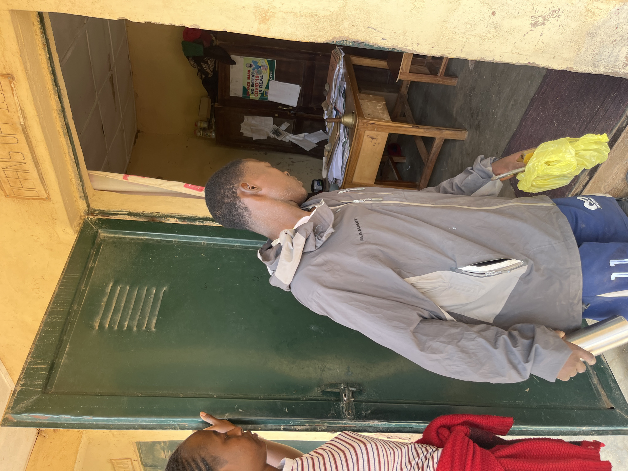 A man wearing a grey jacket and blue shorts stands in a doorway, holding a silver water bottle in his left hand and a yellow plastic bag in his right. He is facing into a room that appears to be an office, with a wooden desk cluttered with papers and a small bell. Behind the desk is a wooden cabinet with several items on top and papers attached to its doors. A sign with text about COVID-19 is visible on the cabinet door. A woman wearing a striped shirt and a red scarf is partially visible on the left side of the doorway, leaning against the green door. The exterior of the building is painted yellow, and the doorway is framed by the yellow wall. Above the doorway, there is text on the wall that reads "OFFICE". This scene appears to be located in Bukuru, Nigeria.