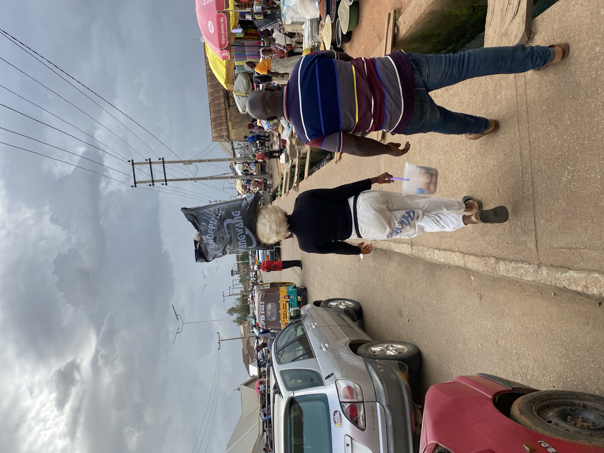 This is a forensic, matter-of-fact description of an outdoor market scene in Bukuru, Nigeria. The image depicts a street with a paved walkway on the right side, bordered by a narrow, open channel. The sky is overcast with heavy, gray clouds.
On the walkway, a woman is walking away from the camera, carrying a large black plastic bag labeled "SHOPPING" on her head. She has blonde, curly hair and is wearing a black long-sleeved shirt, white pants with a blue design, and dark sandals. She is also carrying a clear plastic bucket containing what appear to be eggs.
Walking beside and slightly behind her is a man, also moving away from the camera. He is wearing a striped short-sleeved shirt in shades of purple, blue, white, and yellow, blue jeans, and brown sandals.
The street itself is a dirt road with some loose gravel. Several vehicles are visible, including a silver SUV on the left with an "OPEN DOORS" sticker on the rear passenger door, and a red car partially visible in the lower left corner. Further down the street, a yellow and green auto-rickshaw (a "Keke") with the license plate "PTA 1265" is stationary. A man in a red uniform stands near the auto-rickshaw.
The market area is to the right of the walkway. Stalls are set up with colorful umbrellas, including a pink "itel" branded umbrella with the tagline "Enjoy better life." Various goods are displayed, including sacks of grain, bowls of what appear to be spices or grains, and other items. Several people are visible in the market area, including shoppers and vendors. Overhead power lines and utility poles are present in the background, crossing the street. The overall impression is that of a busy, active marketplace under a cloudy sky.