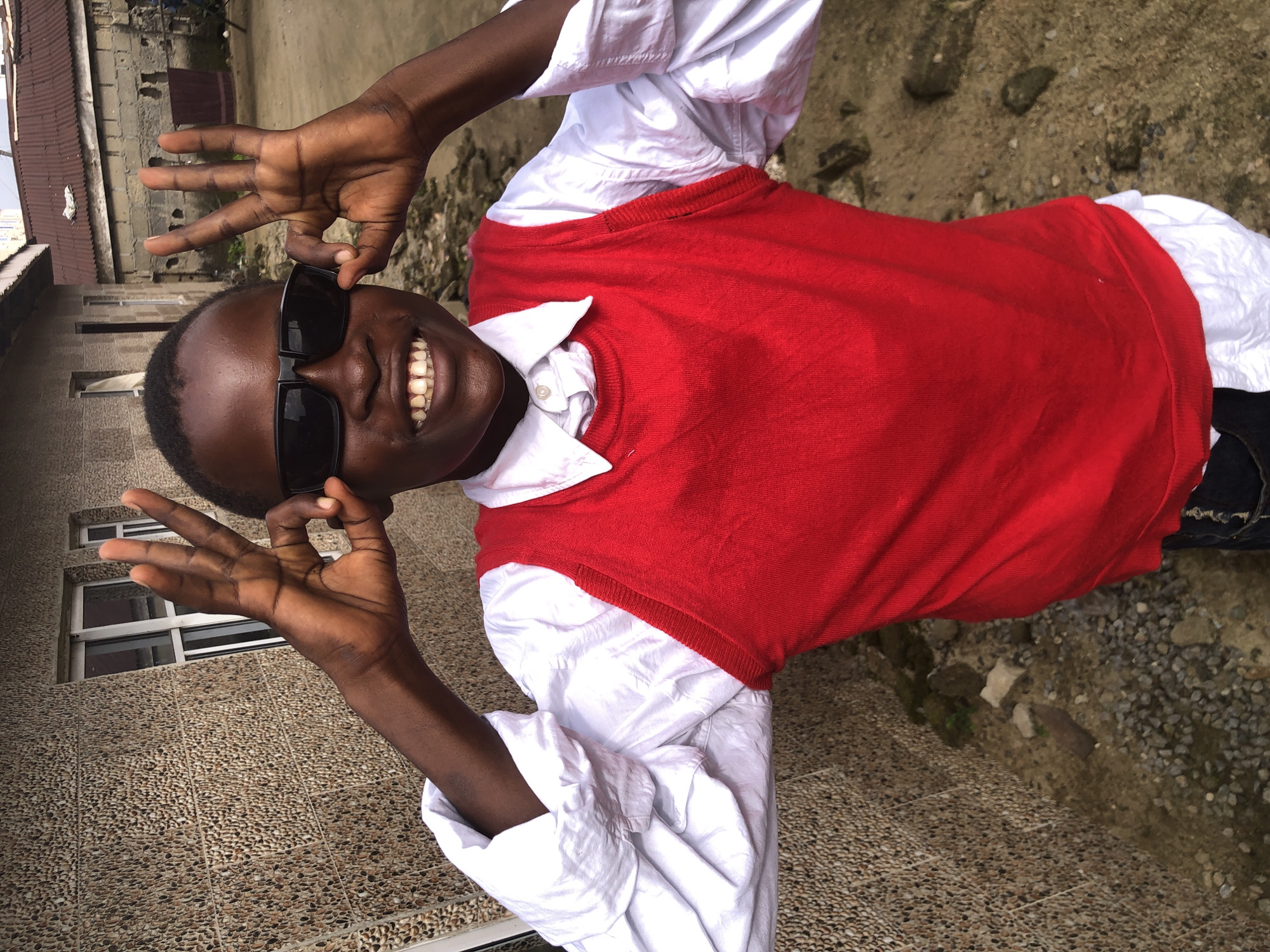 A medium shot shows a young Black boy wearing sunglasses and a red sweater vest over a white collared shirt. He is standing in what appears to be an outdoor setting in Ado, Nigeria, with a textured, stone-like building wall on the left and other buildings and a dirt/gravel ground in the background. The boy is holding up both hands, with his index fingers and thumbs forming circles, and his other fingers extended upwards, creating a peace sign or "okay" gesture near his eyes. His mouth is open in a wide smile, revealing his teeth. The lighting suggests it is daytime.