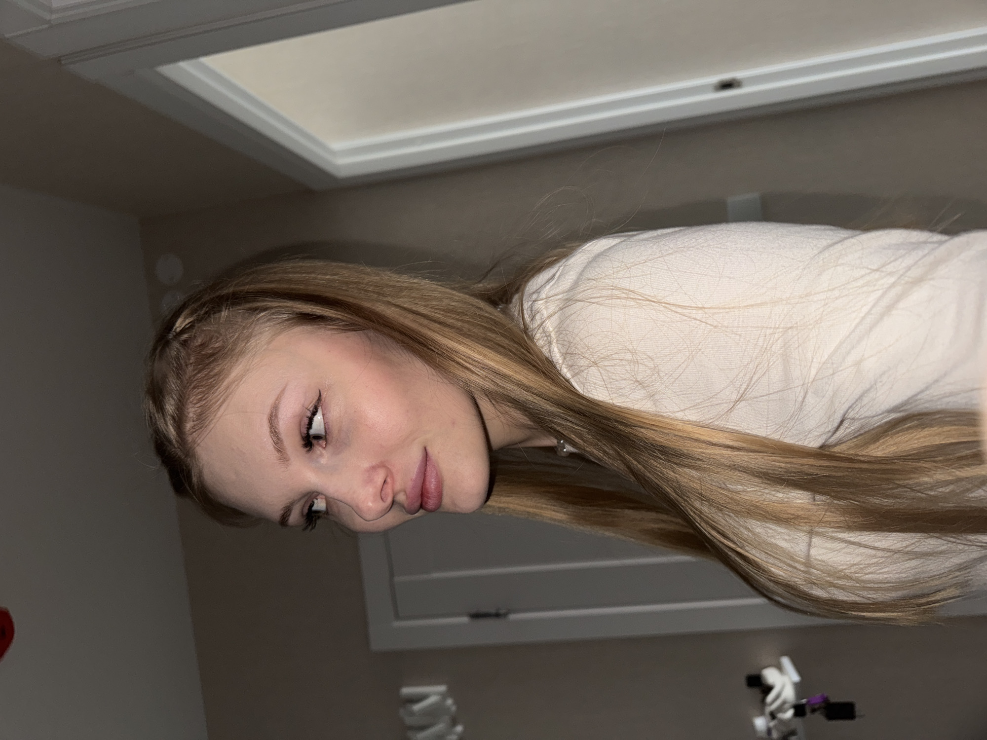 A young woman with long blonde hair, styled with a small braid at the crown, is positioned in profile facing left. She is wearing a light-colored, long-sleeved shirt. Her makeup includes winged eyeliner. She appears to be indoors, in what looks like a hallway. There is a white doorframe to her right, and a lighter colored door behind her to the left. The walls are a neutral beige color. The lighting is dim. No specific location in Perm, Russia can be identified from the image.