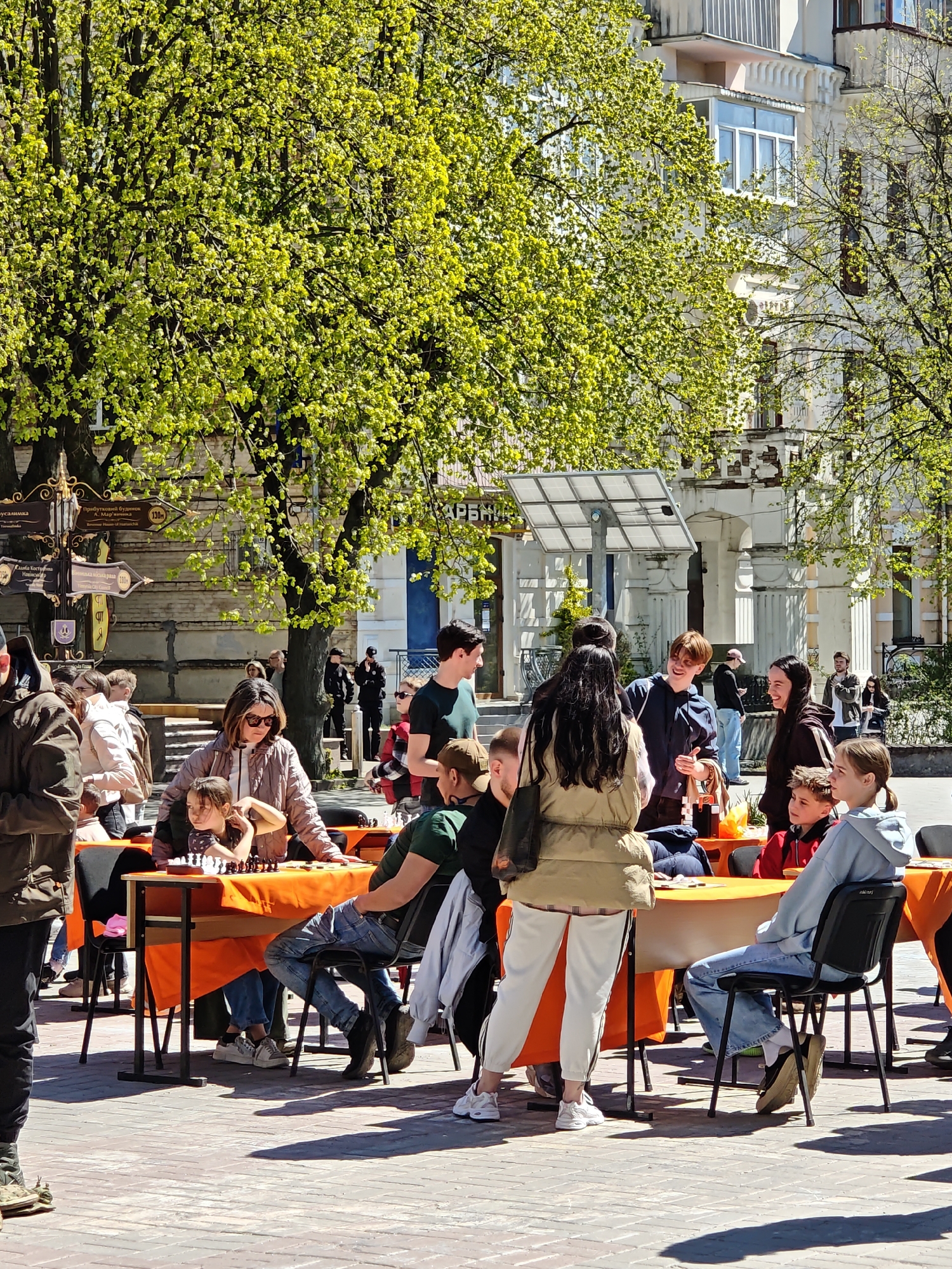 The image captures an outdoor scene in Vinnytsia, Ukraine, with a group of people gathered around tables covered in orange tablecloths. Some individuals are seated and appear to be engaged in playing chess, while others are standing and interacting. The environment includes buildings in the background and trees with lush green foliage. A signpost with multiple signs pointing in different directions is visible, indicating various locations within the city. The people's clothing suggests it is a casual, daytime event. The overall atmosphere suggests a social gathering or outdoor activity in a public space.