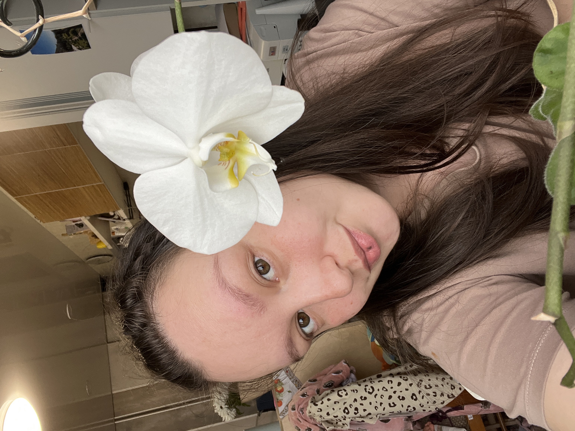 A selfie-style close-up of a woman with long, dark brown hair. She is fair-skinned with brown eyes. A large white orchid is positioned near her head, seemingly placed in her hair. She is wearing a light brown top. The background includes glimpses of a room with various items, including bedding, a lamp, and a piece of furniture. The location appears to be indoors.