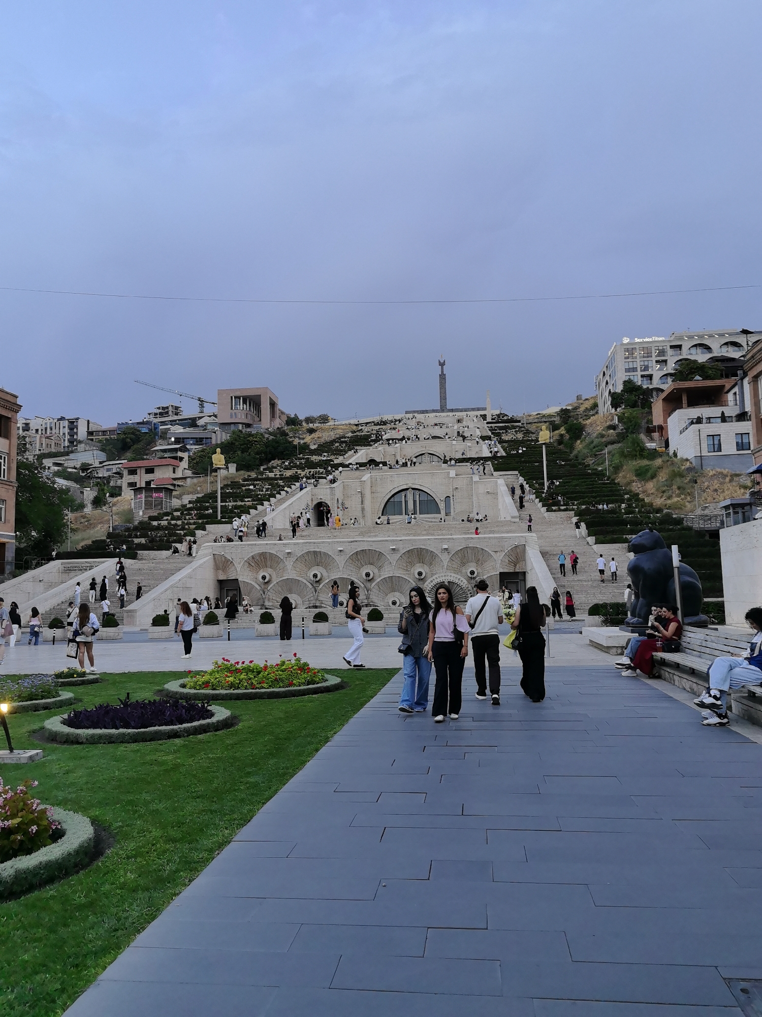 This image depicts the Cascade Monument in Yerevan, Armenia. The monument is a giant staircase adorned with fountains, gardens, and sculptures, leading up a hillside. In the foreground, several people are walking along a paved pathway, with manicured green lawns and flowerbeds on their left. A large black sculpture of a lion is visible on the right. Further up the cascade, many people are seen ascending the stairs, with buildings and trees dotting the hillside. The sky is overcast, suggesting it is either early morning or late evening.