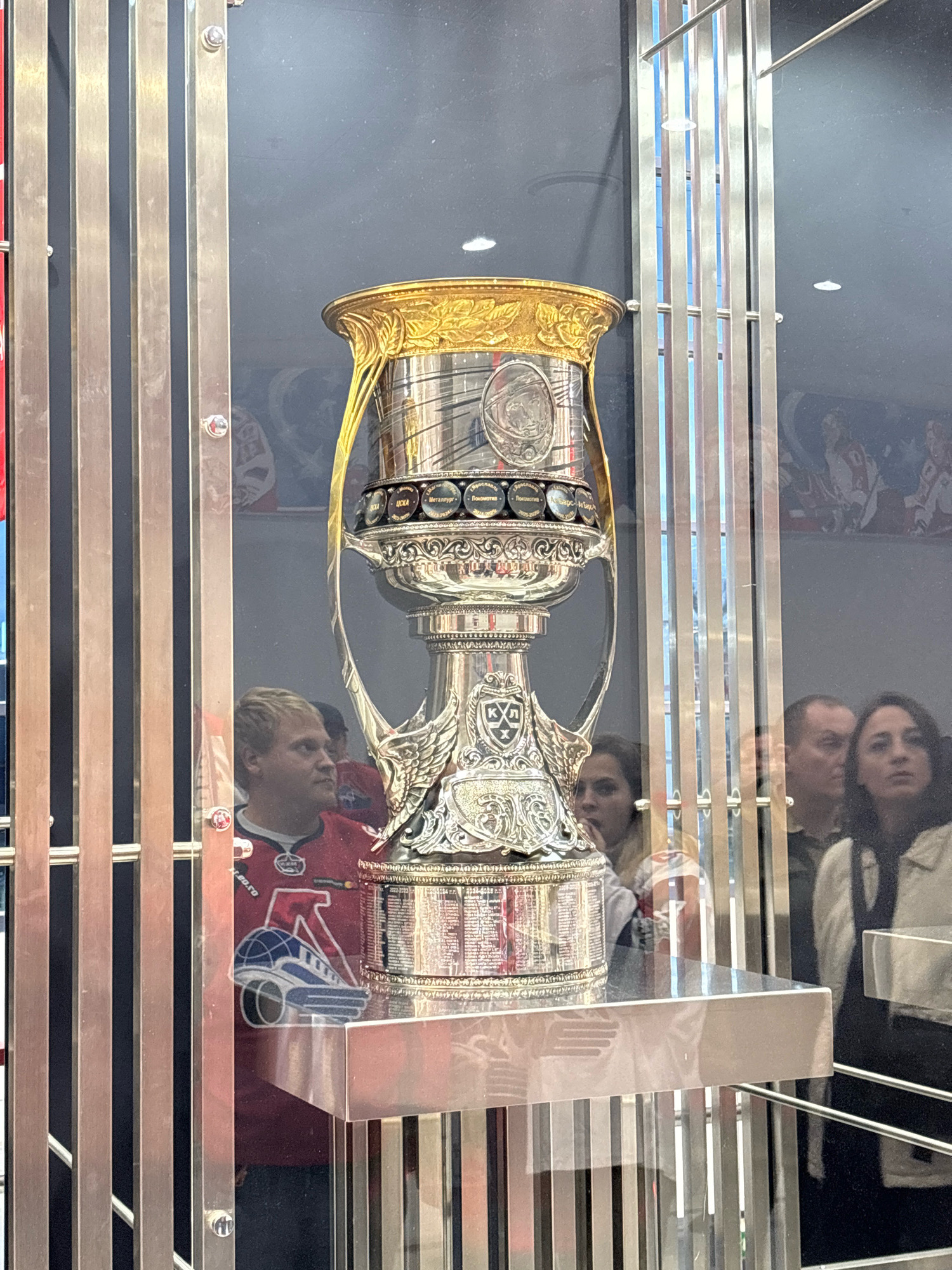In Rusanovo, Russia, a large, ornate silver and gold trophy is displayed behind a glass enclosure. The trophy features intricate detailing, including engraved medallions around its base, wings on its handles, and a golden rim. Several people are visible looking at the trophy through the glass. On the left, a person wearing a red hockey jersey with a logo is partially visible. To the right, a group of individuals are observing the trophy. The environment appears to be a museum or exhibition hall, with a dark background and reflective surfaces.