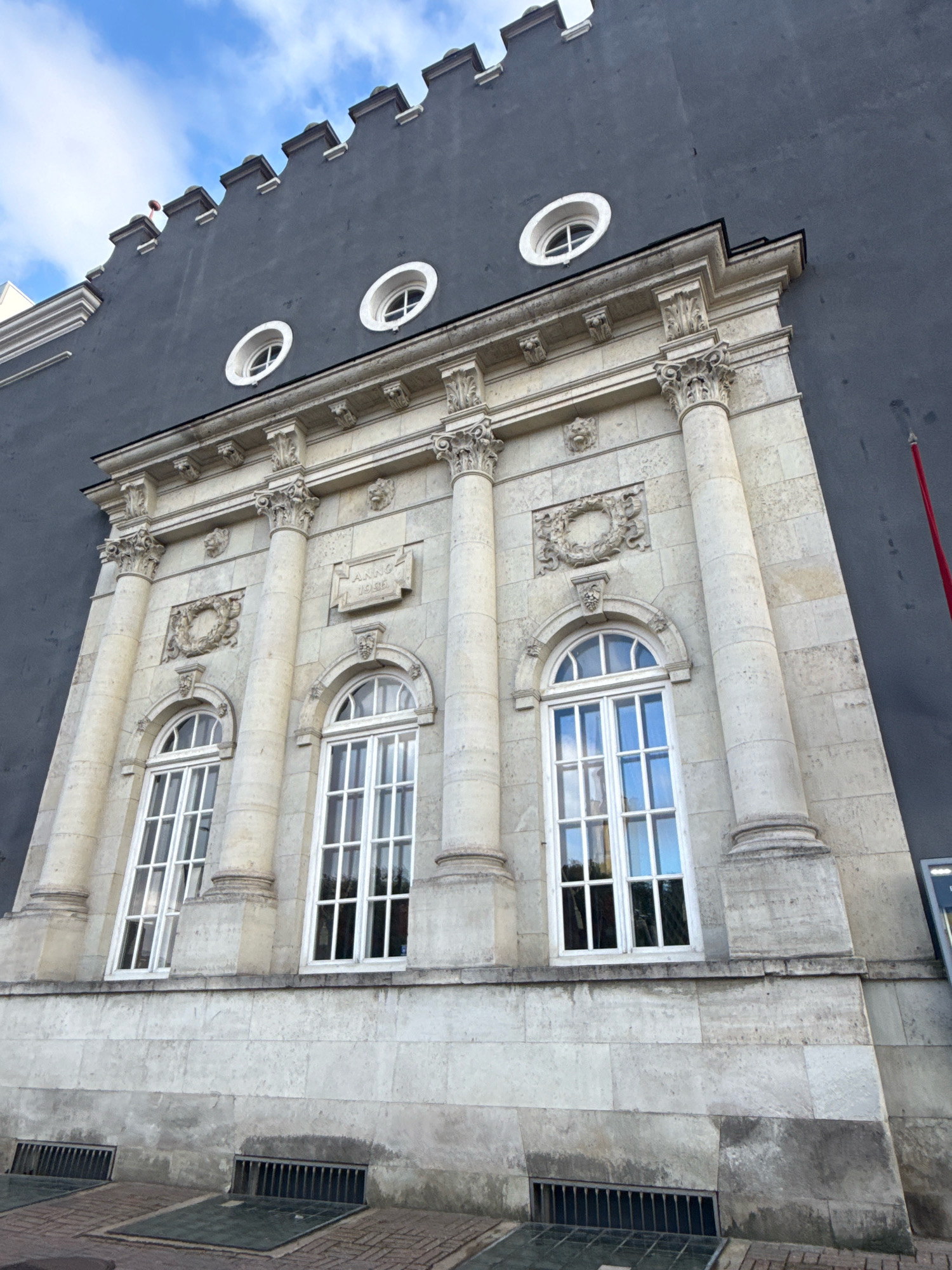 The image shows the facade of a building in Tallinn, Estonia. The building has a gray, textured upper wall with a crenellated top. Below this, there is a section of pale stone construction with classical architectural elements. This stone section features three tall, arched windows with white frames and multiple panes. The windows are separated by fluted columns that rise from a stone base. Above the windows, there are decorative circular wreaths and small carved lion heads. A rectangular plaque with the inscription "ANNO 1936" is visible on the stone facade. In the upper part of the gray wall, there are three circular windows with white surrounds. The lower part of the building shows a stone wall with two horizontal barred vents and a tiled pavement in front of it. The sky above is blue with scattered white clouds.