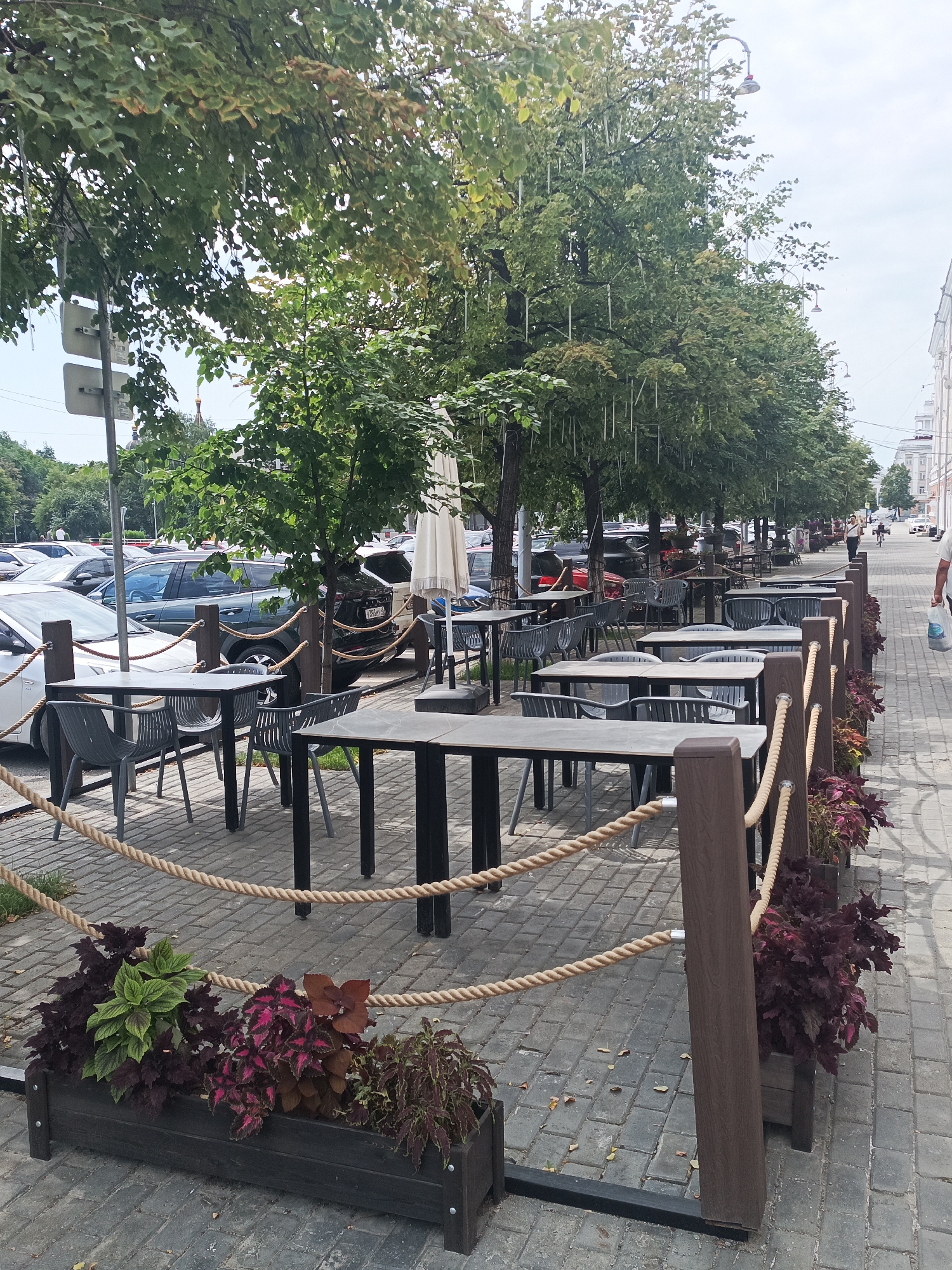 The image shows an outdoor seating area of a cafe or restaurant, likely located in Kurgan, Russia. The seating area is set up along a paved sidewalk lined with trees. There are numerous tables and chairs, suggesting a busy establishment. The tables are dark-colored with metal frames, and the chairs are a light gray plastic. A rope barrier with wooden posts separates the seating area from the sidewalk and the street. Beyond the seating area, cars are parked along the street, indicating an urban setting. In the background, there are buildings and a few people walking, suggesting a public space. The overall atmosphere is one of an active street scene.
