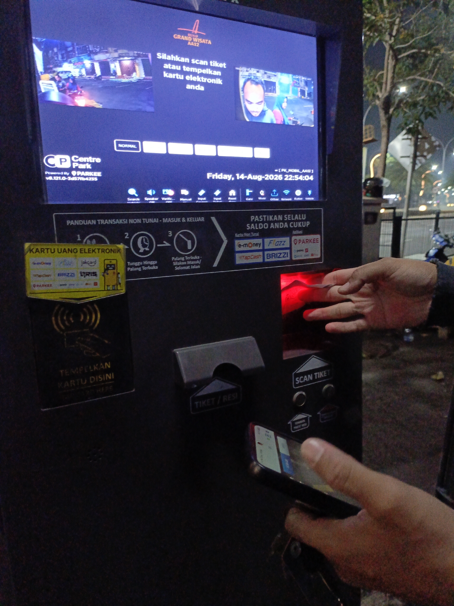 An automatic ticket and payment machine is shown at night in Lambangjaya, Indonesia. The machine has a touchscreen display showing a video and text in Indonesian. Below the screen, there are instructions for using electronic money cards, with logos for various payment providers. A person's hand is shown interacting with the machine, holding a smartphone near a red scanner and also holding an electronic card to a reader. There are also physical card slots and a ticket dispenser on the machine. The surrounding environment appears to be outdoors, with a tree and streetlights visible in the background.