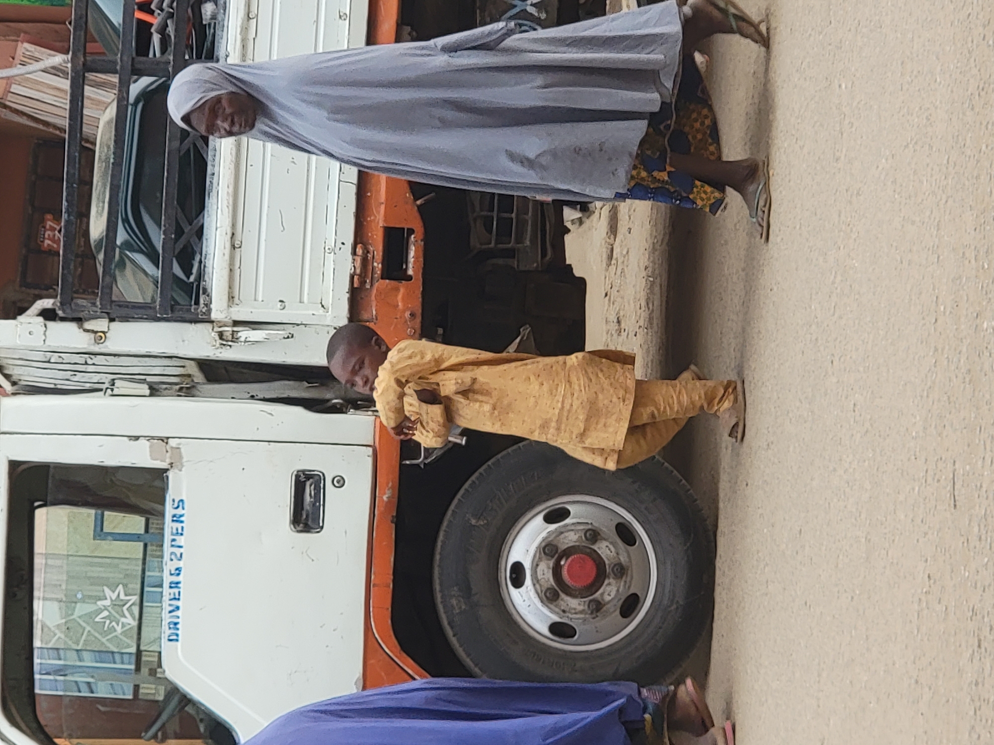 The image, taken in what appears to be a street scene, features two individuals standing next to a parked truck. On the left side of the frame, a white and orange truck dominates, showing its cab, a front wheel, and a portion of its chassis.
An adult, likely female, is positioned towards the upper right of the truck, wearing a light grey head covering that drapes over a patterned dress featuring blue, yellow, and black designs. Her feet are visible, clad in light-colored flip-flops. Further down and slightly in front of her, closer to the truck's front wheel, a child stands, dressed in a light yellow or beige long-sleeved garment. The child appears to be peeking or looking towards the viewer from behind the truck's mudguard. Both individuals are looking generally in the direction of the camera.
The background includes indistinct structures, possibly buildings or parts of other vehicles. The ground is a light, dusty color, suggesting an unpaved or lightly paved surface. The lighting is bright but diffused, indicating daytime, possibly under an overcast sky, as there are no harsh shadows.
Visible text on the white door of the truck reads "DRIVERS 2 PERS" in blue lettering. A faint red plate with the number "737" is partially visible behind the truck's cab. The overall setting depicts a candid moment in an everyday street environment.