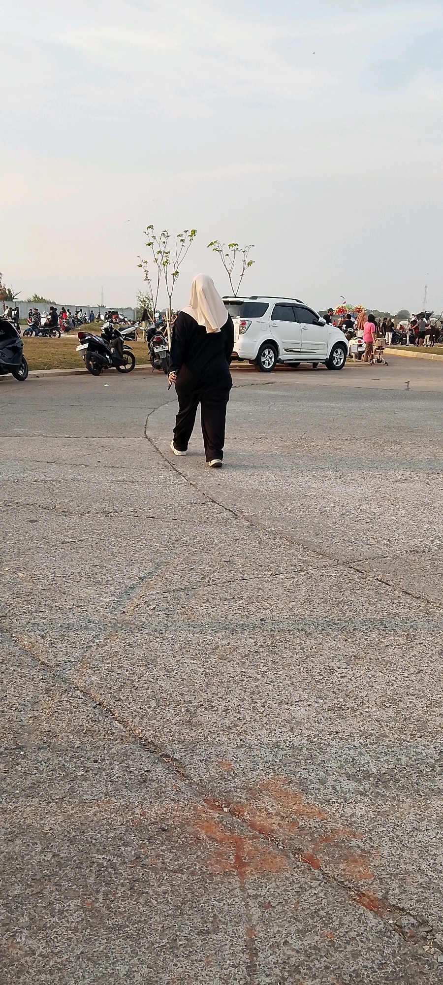 A woman wearing a light-colored hijab and a black long-sleeved outfit walks away from the camera on a paved surface. Behind her, several motorcycles are parked. Further in the background, a white SUV is parked, and a group of people are visible. The scene appears to be an outdoor parking area or gathering space, potentially in a city like Satriamekar, Indonesia, with a bright sky and sparse vegetation. The ground shows some cracks and reddish stains.