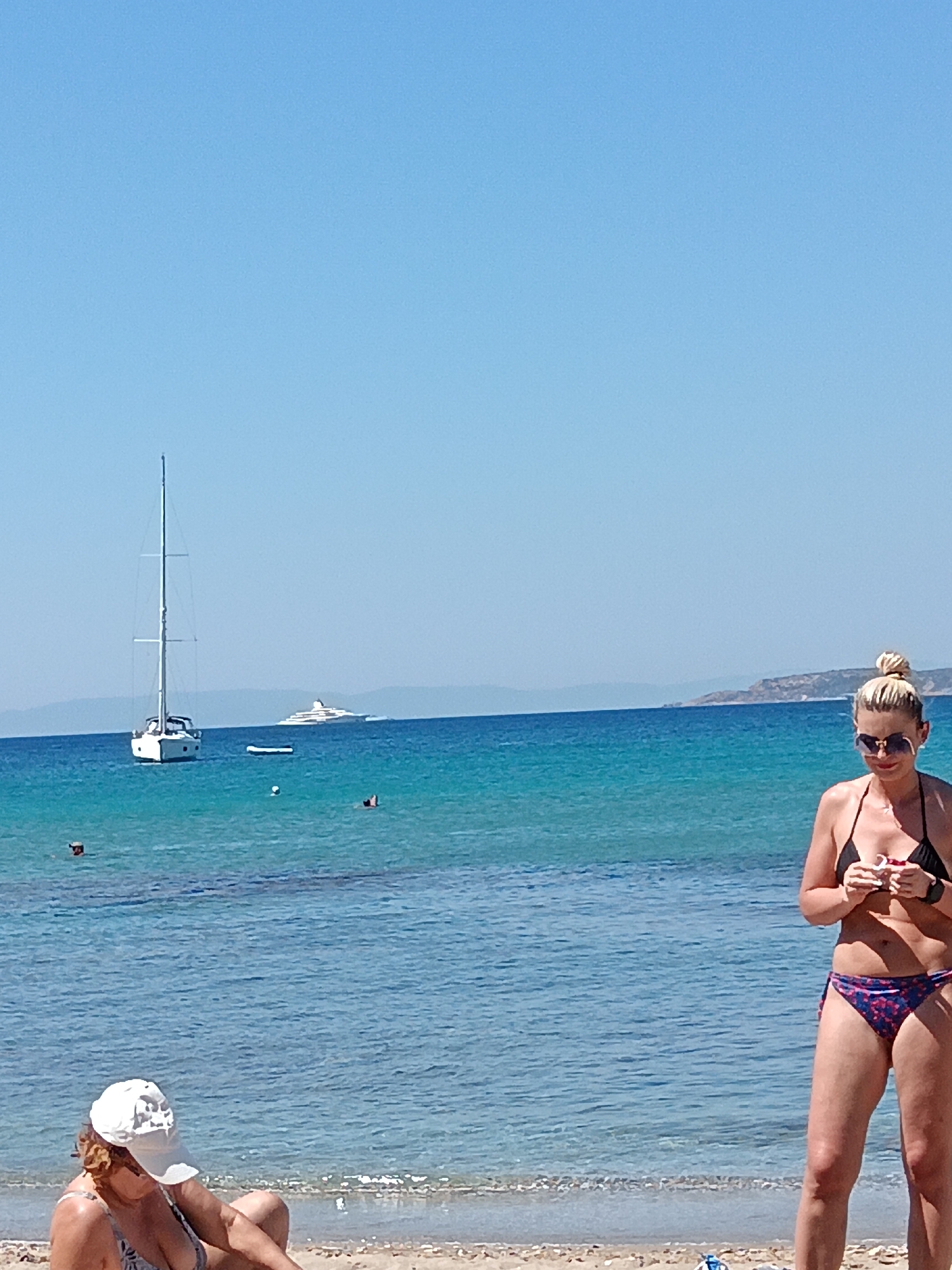 In the city of Agia Marina, Greece, on a sunny day, people are enjoying a beach scene with the turquoise Aegean Sea. In the foreground, a woman with blonde hair in a bun, wearing sunglasses and a black bikini, stands on the sandy beach. She is holding something in her hands. To her left, another person wearing a white cap and a patterned bikini is partially visible, sitting on the beach.
In the water, a white sailboat with tall masts is anchored. Further in the distance, a large white yacht is visible on the horizon. There are a few other people in the water, some swimming or wading. The sky is clear and blue, and the sea is calm and inviting. The background features a hazy, mountainous coastline.