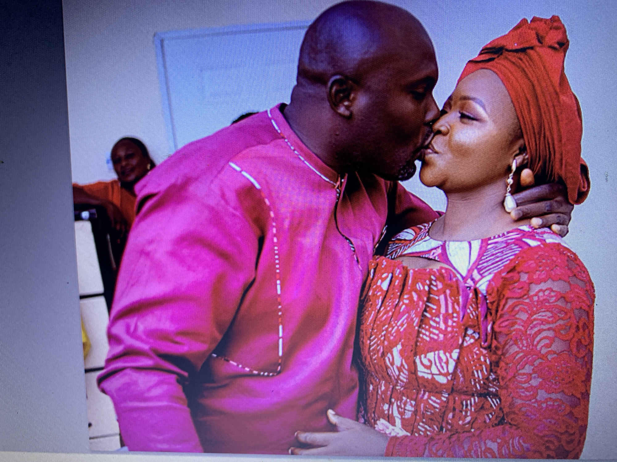A medium shot shows a couple in a close embrace, with the man leaning in to kiss the woman. The man, with a shaved head, wears a vibrant fuchsia kaftan with white and red bead detailing. The woman, adorned in a matching red headwrap and earrings, is dressed in a red and white patterned dress with lace accents. Her eyes are closed, and the man's arm is around her neck. In the background, a blurred figure wearing an orange top is visible, seated. The setting appears to be indoors, with a light-colored wall and a light blue door or partition visible behind the couple. The overall scene conveys an intimate and celebratory moment.