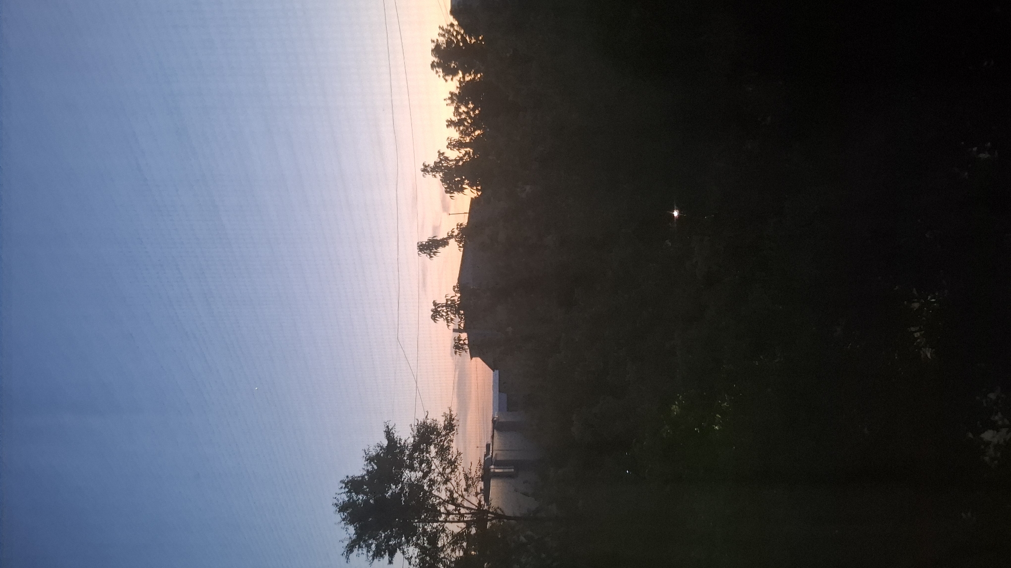 The image captures a scene at dusk, with the sky displaying a gradient of blue to soft pink and orange hues. Silhouetted against this twilight sky are trees and a rugged, elevated landscape, possibly a hill or mountains. In the foreground, a solitary tree with dark foliage stands out. The right side of the image is predominantly dark, suggesting it's transitioning into night, with the shapes of trees and foliage barely discernible. A small, bright light, possibly a distant lamp or star, is visible in the dark section. There are also power lines visible in the sky on the left side of the image. The overall mood is serene and tranquil, indicative of the end of the day. No people or text are visible in the image. The location context suggests this is Shymkent, Kazakhstan.