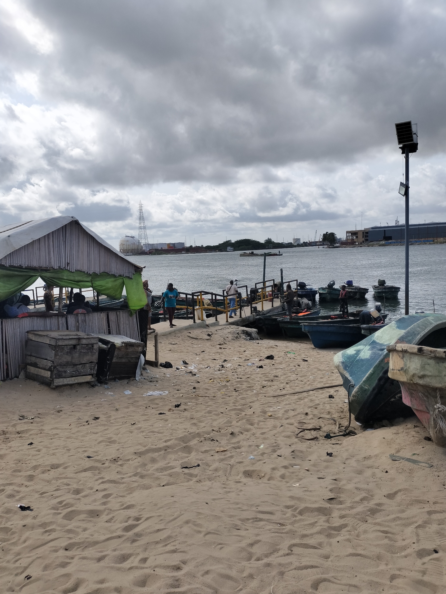 The image depicts a waterfront scene in Lagos, Nigeria. The foreground is dominated by a sandy beach littered with debris. There is a makeshift structure on the left, seemingly a shaded shelter, constructed with corrugated metal sheets and green tarpaulin. Several people are inside the shelter. To the right, a small pier extends into the water, with several small fishing boats docked alongside. People are walking on the pier and working on the boats. In the background, across the water, are industrial buildings and structures, including a tall electrical tower. A tall lamppost stands on the right. The sky is overcast with dark, heavy clouds.