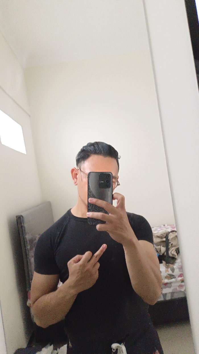 A man is taking a selfie in a mirror. He is wearing a black t-shirt and has his middle finger extended. He is holding a black smartphone in his right hand and is looking at the camera. The room has white walls and there is a bed with a headboard in the background. This image was taken in Magelang, Indonesia at the coordinates: latitude -7.479, longitude 110.222. The image is taken from the waist up. The lighting in the room is bright, and the man's face is illuminated. The background of the image is out of focus, but a bed and a floral patterned blanket can be seen. The man's left hand is obscured from view. The room is cluttered with personal belongings.