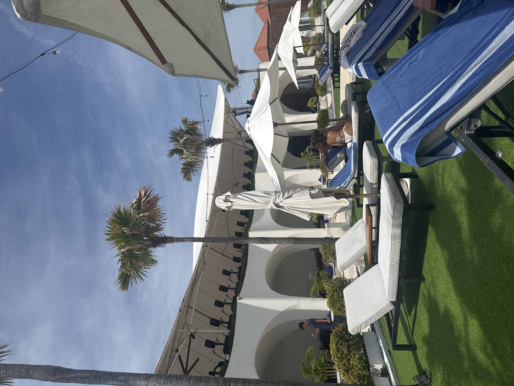 The image depicts an outdoor setting, potentially a beach club or resort in North Kuta, Indonesia, given the presence of tropical vegetation, palm trees, and architectural styles common to the region. The scene is dominated by white structures featuring arched openings, possibly a building or a covered patio area. Lounge chairs are arranged on what appears to be artificial grass. Several large white umbrellas provide shade. A woman in swimwear is seen reclining on one of the lounge chairs. A man wearing a blue shirt is walking nearby.