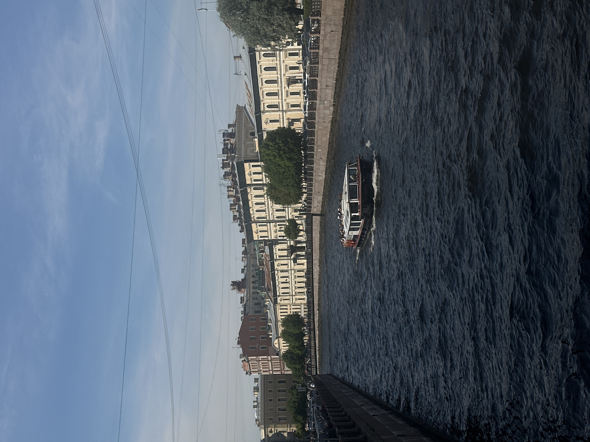 An eye-level shot captures a boat navigating a dark, choppy river that flows between two stone embankments in a city. The embankments are lined with trees and grand, multi-story buildings with cream-colored facades and ornate detailing. A clear blue sky with thin clouds stretches above, with visible power lines crossing the frame. The boat, with a white upper deck and dark hull, is moving away from the viewer, and several people can be seen on its deck. The left embankment is lined with parked cars, and a few trees are visible along the right embankment. The scene appears to be a sunny day in an urban environment with historical architecture.