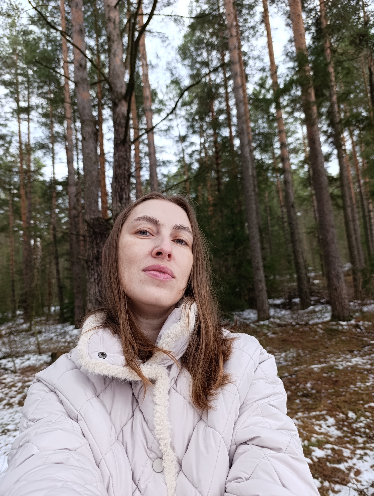 A woman is standing in a sparse pine forest. Patches of snow are visible on the forest floor. She is wearing a light-colored, quilted jacket with a fur-trimmed collar. The location is Chaikovsky, Russia.