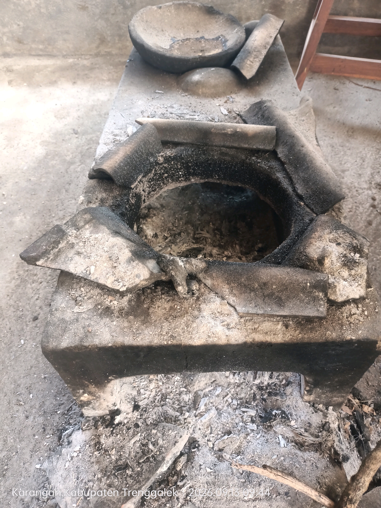 The image shows a close-up, overhead view of a traditional outdoor cooking stove. The stove is constructed from dark, weathered bricks and appears to be old and well-used, with visible soot and ash accumulated in and around the fire pit. The fire pit itself is circular, and remnants of burnt wood and ash are present within it. Around the opening of the fire pit, there are several broken pieces of what appear to be clay or ceramic material, possibly part of the original structure or used as a grate. In the background, there is a blurred view of a cement or dirt floor and a portion of what looks like wooden furniture. The bottom right corner of the image displays a watermark indicating "Karangan, Kabupaten Trenggalek, 2026.09.13 09:14". The lighting suggests it is daytime. There are no people or animals visible in the image.