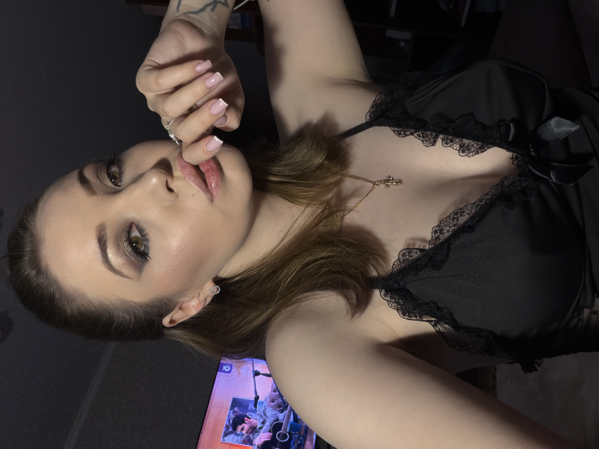 A woman is shown in a close-up, head-and-shoulders shot, looking towards the camera. She has on makeup, including eyeshadow, eyeliner, and lipstick. She is wearing a black, lacy top, a gold necklace with a cross pendant, and a ring on her finger. Her hand is positioned near her mouth. In the background, a television screen displays an image of a man playing a guitar. The lighting is dim, suggesting an indoor setting at night.
