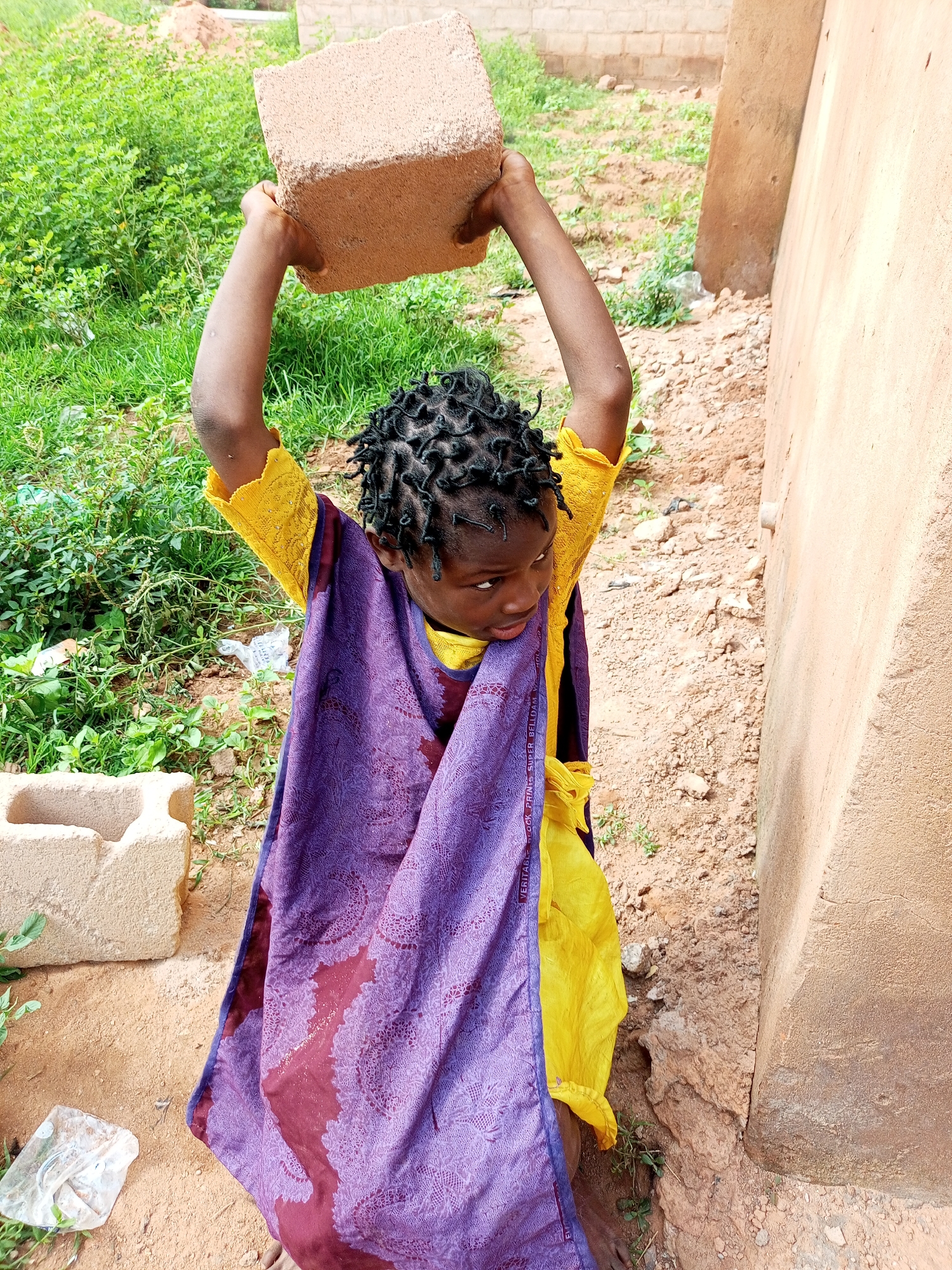A young Black child with dark, coiled hair, wearing a yellow garment and a purple and burgundy patterned cloth draped over it, stands outside a tan building. The child is holding a light brown brick overhead with both hands. The environment consists of dry, sandy ground with some patches of green grass and foliage. In the background, construction materials such as blocks are visible, along with a partially constructed wall. The lighting suggests it is daytime.
