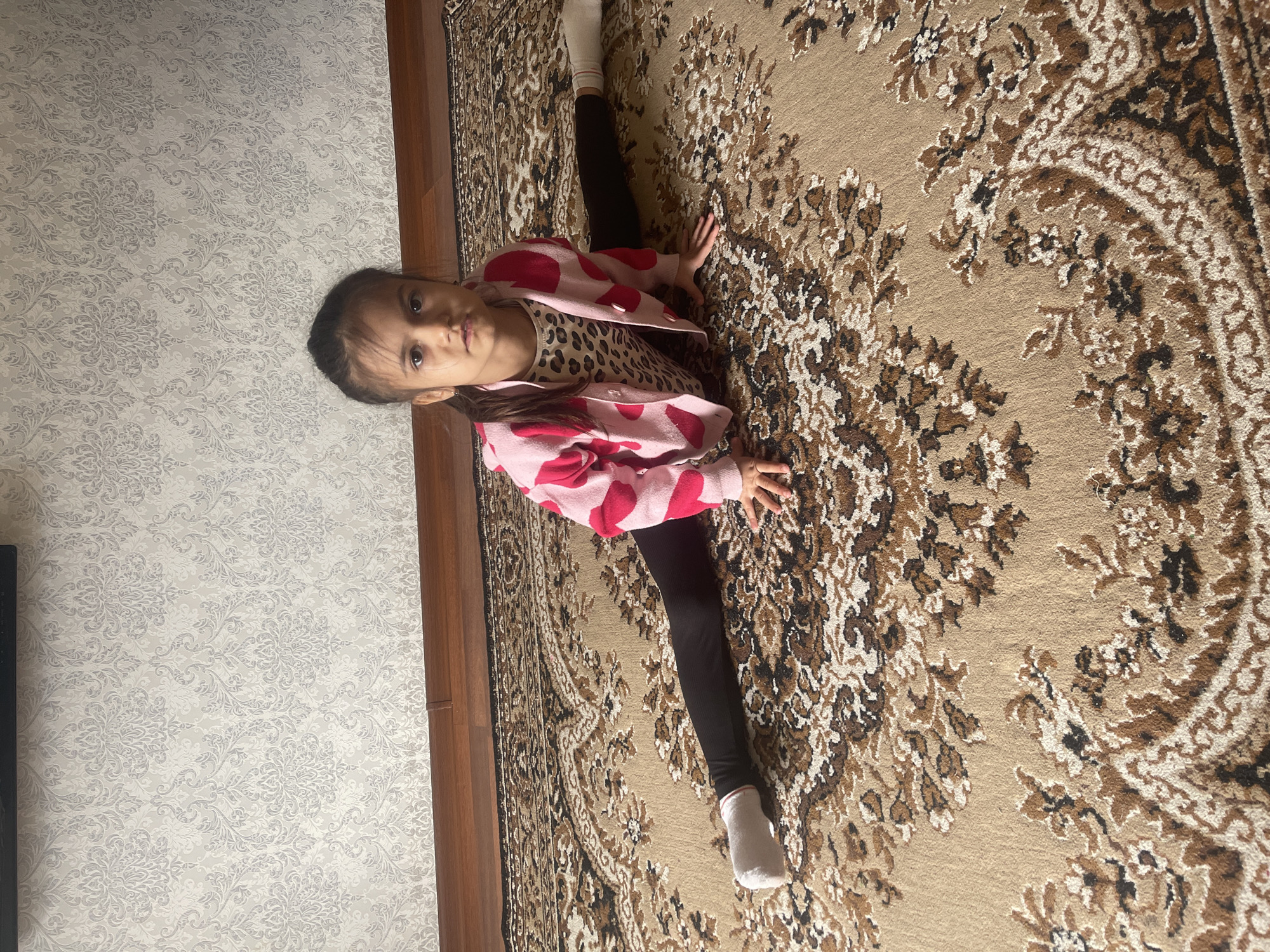 A young girl is captured performing a full splits stretch on a patterned carpet indoors. She is positioned centrally in the frame, wearing a pink cardigan with prominent red, heart-like shapes, over a leopard print top. Her lower body is clad in black leggings and white socks, with her legs fully extended to either side, demonstrating significant flexibility. Her hands are placed on the carpet for support, and she looks directly at the camera with a neutral, focused expression.
The floor is covered by an ornate carpet featuring intricate floral and swirling patterns in shades of beige, brown, white, and black. To the left, a wall is visible, adorned with light grey or off-white wallpaper displaying a subtle, repeating damask or floral motif. A dark wooden border or furniture piece separates the carpeted floor from the wallpapered wall. The indoor setting appears well-lit, suggesting daytime. The scene depicts a moment of physical activity or practice in a residential environment, located in Poltava, Ukraine.