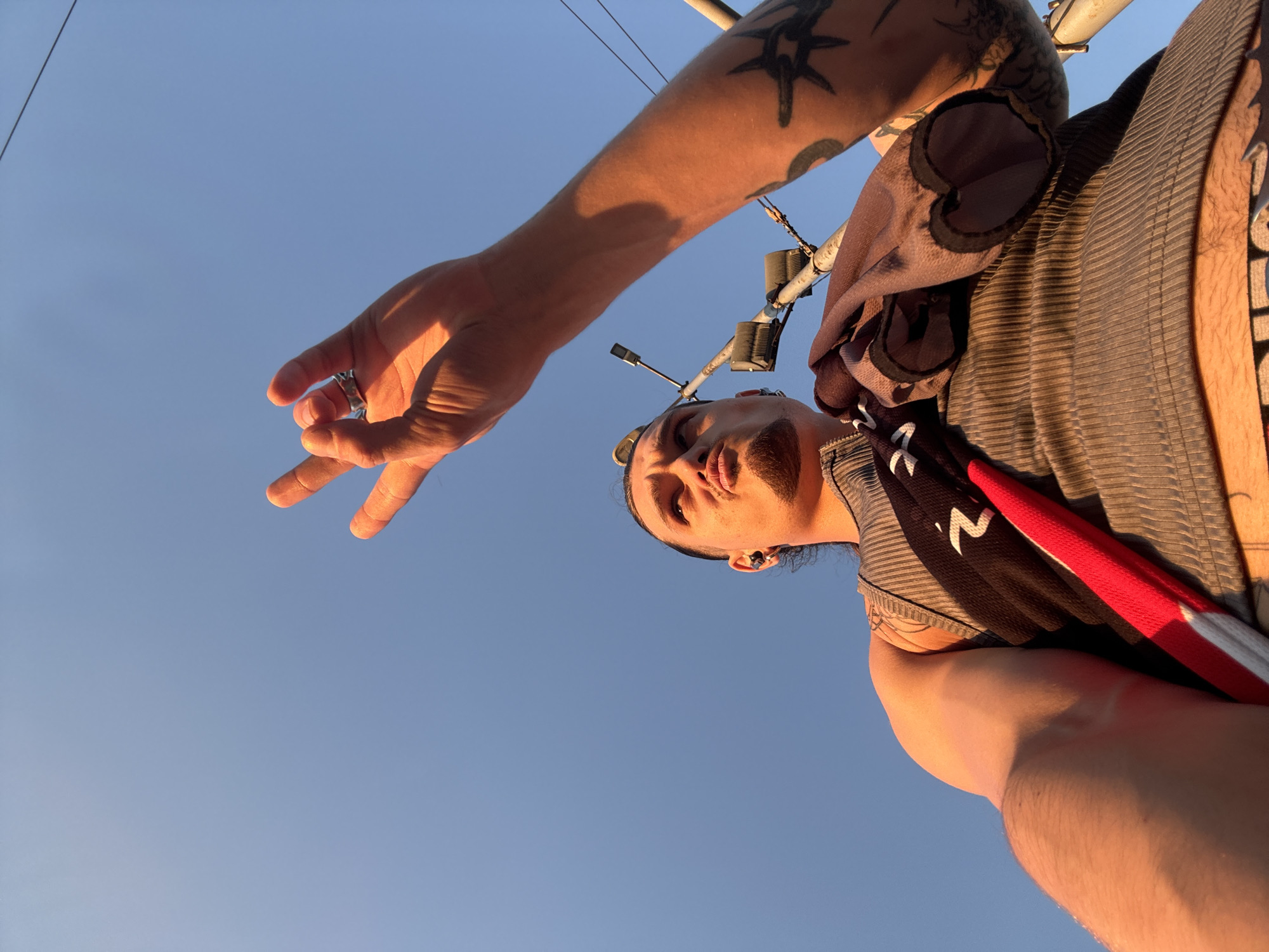 A low-angle, close-up shot captures a person from the chest up, standing outdoors against a clear, bright blue sky bathed in the warm glow of sunset. The individual, whose face is tilted upwards and slightly to the right, sports a goatee and has dark hair pulled back. They are wearing a grey ribbed tank top and have a black and red cloth draped over their shoulder. One arm is raised, with the hand making a gesture with the index and middle fingers extended. A ring adorns the index finger. Visible tattoos are present on the raised arm and shoulder. In the background, a lamppost and power lines are discernible. The lighting suggests it is late afternoon or early evening. The context of Sofia, Bulgaria, is not provided by the image itself and cannot be inferred from the visual information.