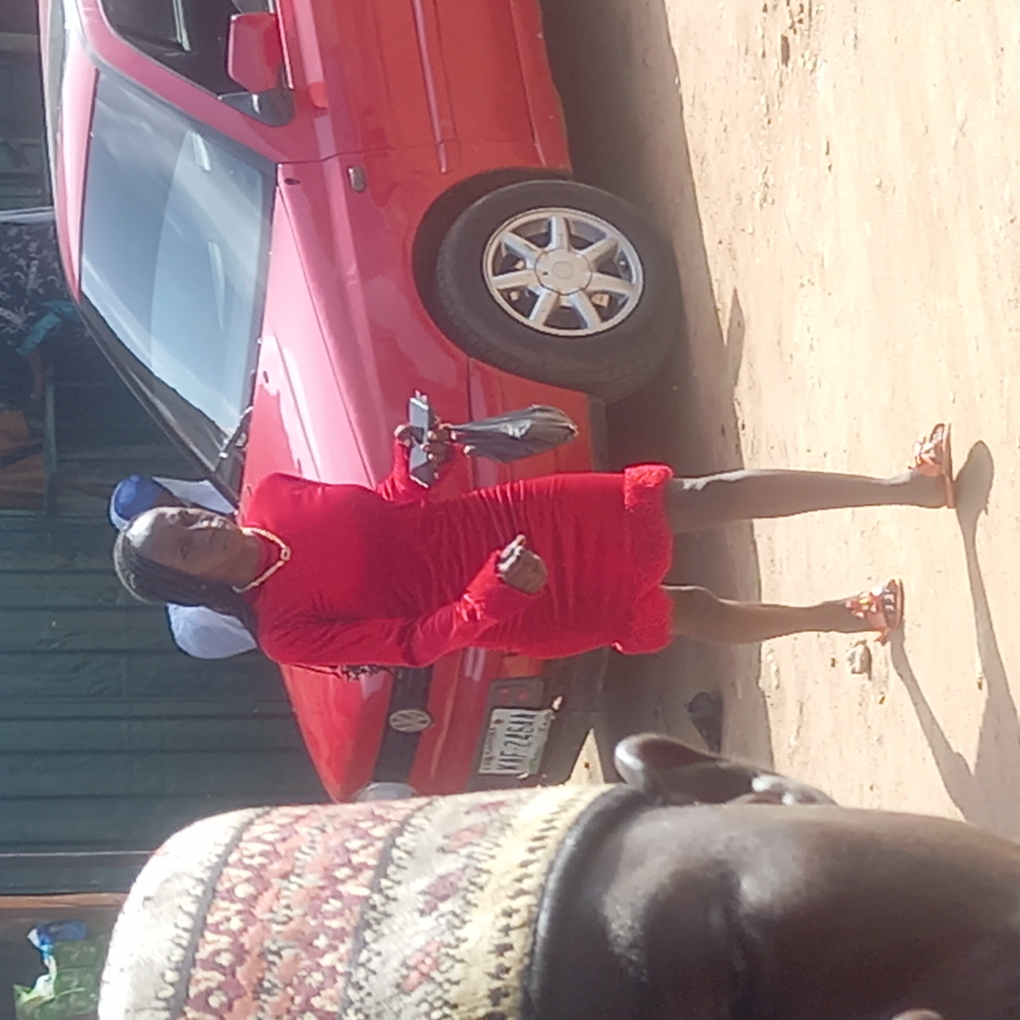 A woman in a red dress stands on a dirt ground next to a red car in Dadi Riba, Nigeria. The woman is holding a cell phone and a black purse. The car has a license plate with the letters "KAF 246AA". There is a partially visible person in the foreground wearing a patterned fabric hat.