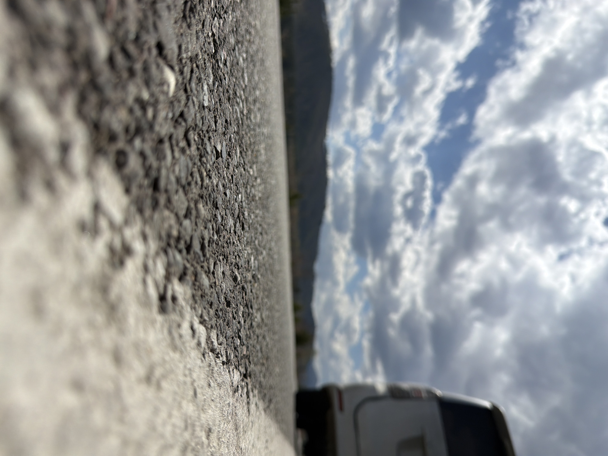 The image, taken at a low angle near a road surface, features a close-up view of asphalt pavement with a white painted road marking. The asphalt texture is coarse. In the background, there is a mountain range and a sky filled with clouds. A vehicle, possibly a camper or van, is partially visible in the lower right corner. Based on the city specified, the location is likely somewhere near Сизая, Russia, but there are no specifics. There are no people shown in the scene.